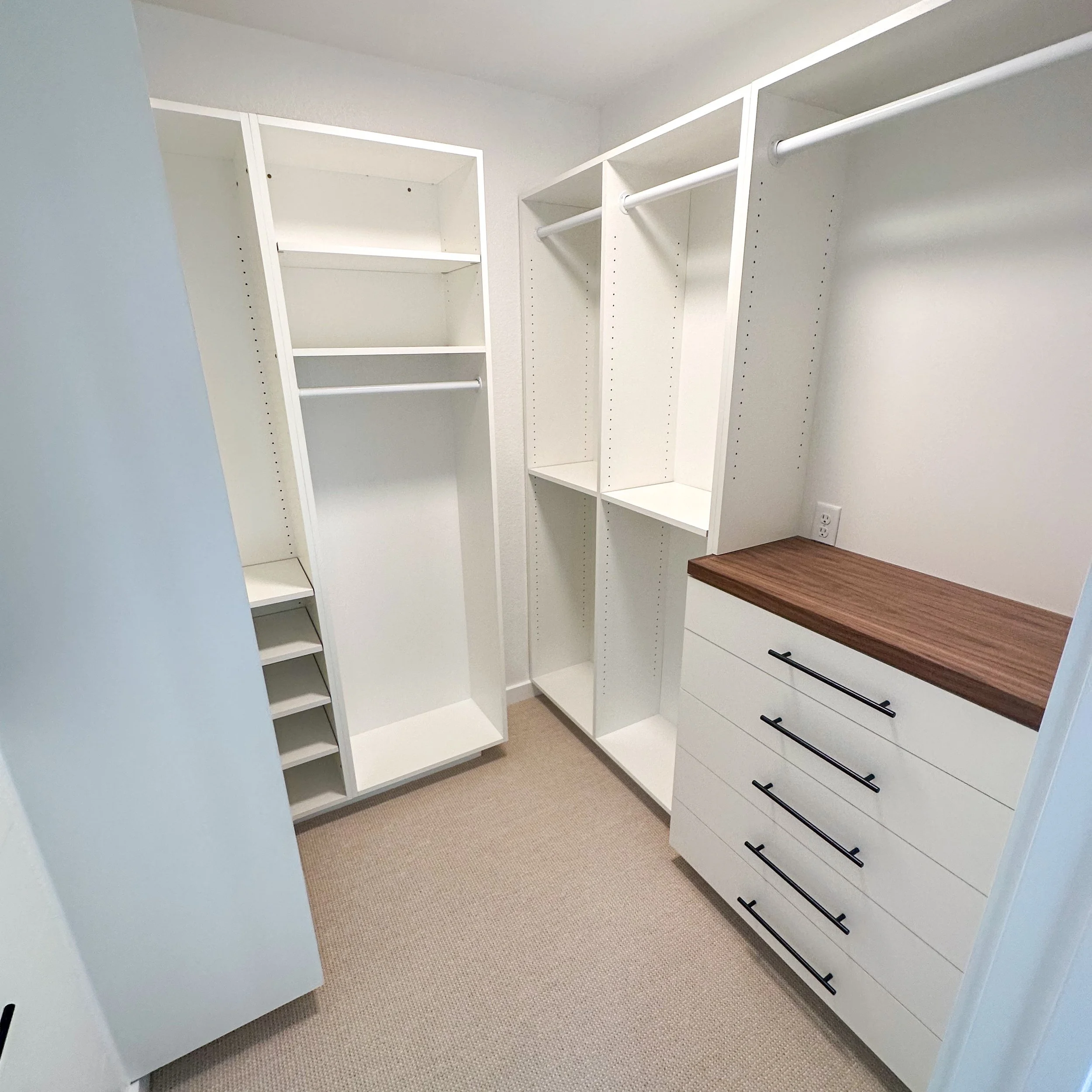 Empty walk-in closet with white shelving units, hanging rods, and a wooden countertop on built-in drawers, beige carpeted floor, and white walls.