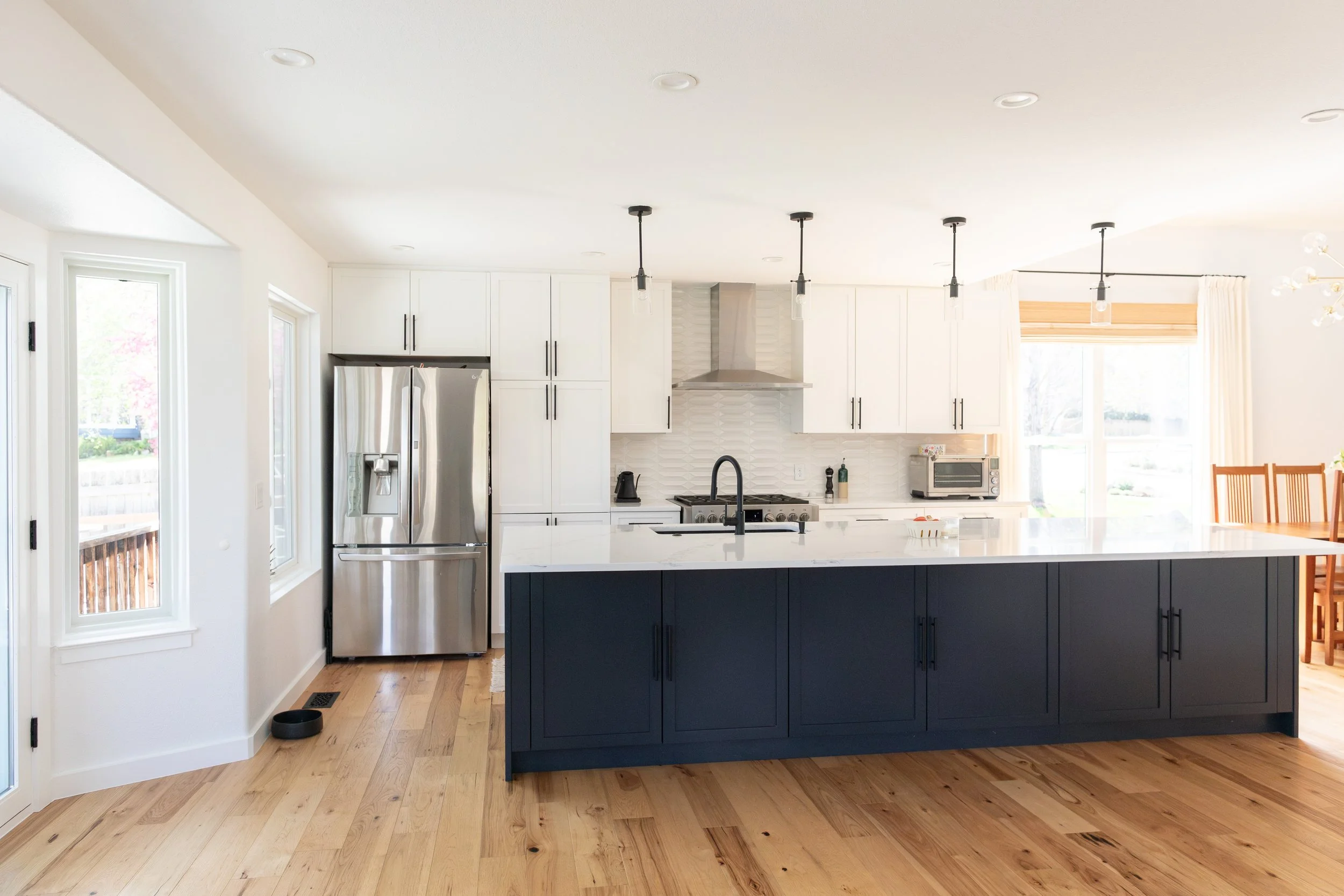 Bright kitchen with white cabinets, stainless steel refrigerator, dark blue island, light wood floors, large windows, and pendant lighting.