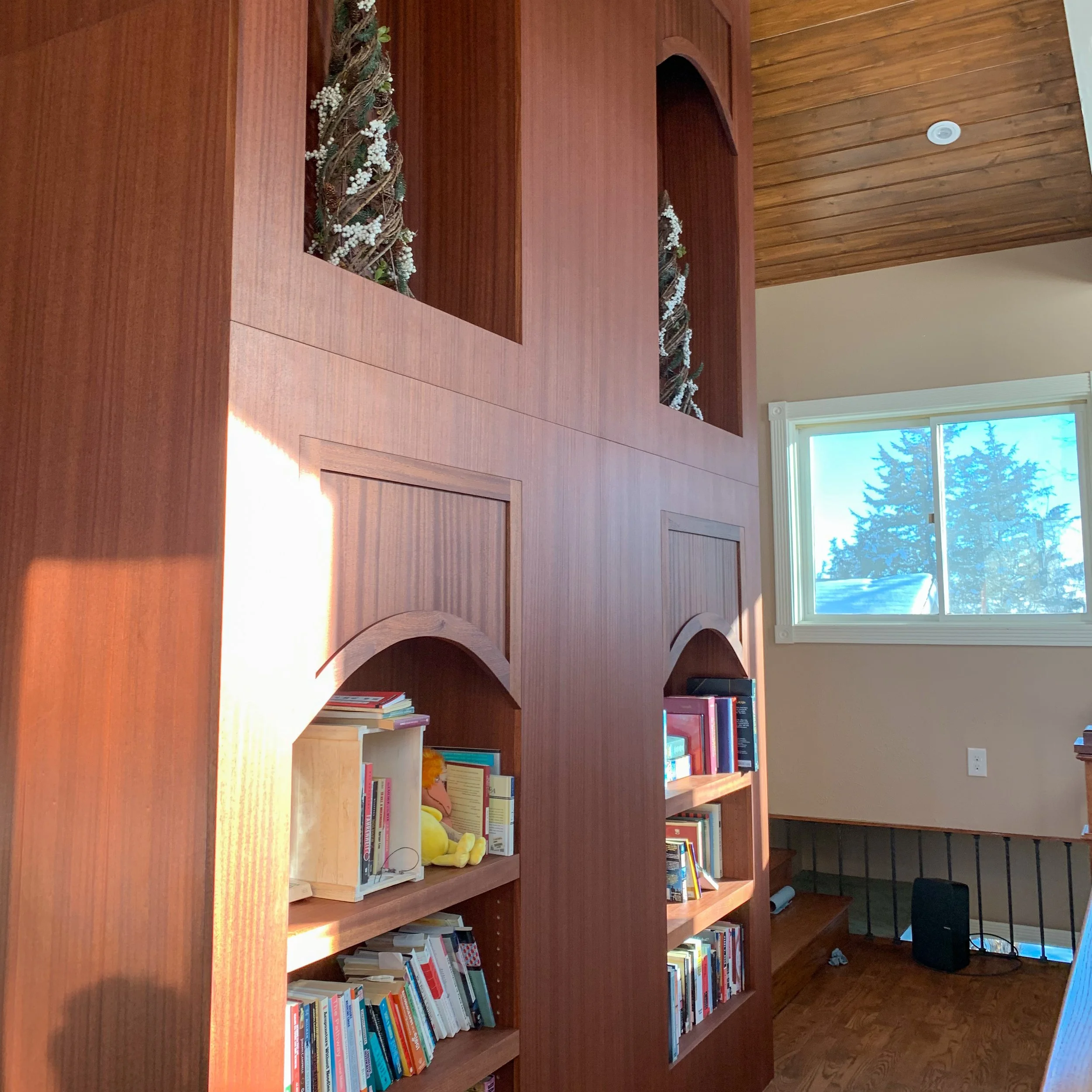 A wooden wall with built-in shelves containing books and a yellow plush toy, near a window with trees outside, in a room with a wooden ceiling and hardwood floor.