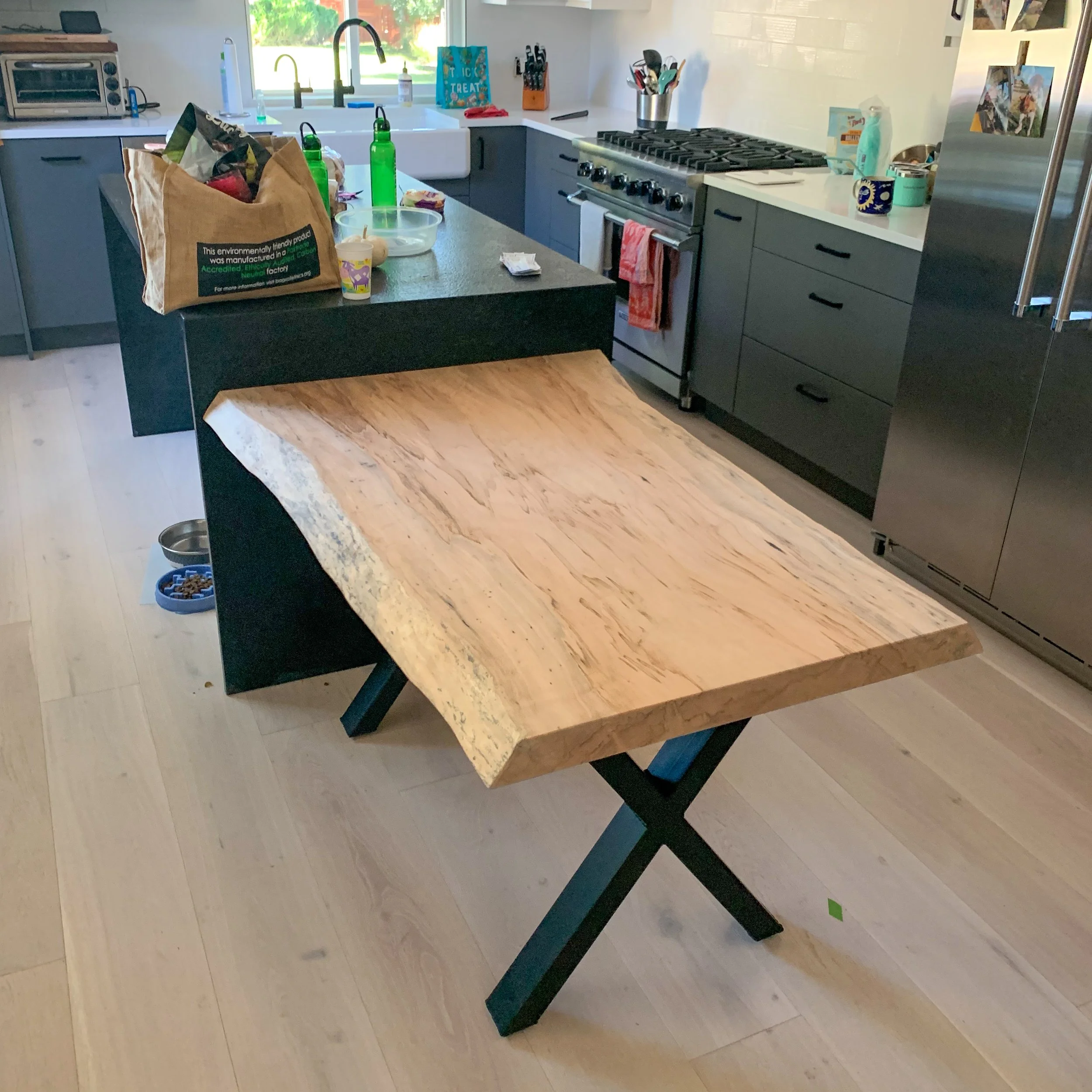 A kitchen with a wooden dining table with black legs, situated near black cabinetry, a gas stove, and a countertop with various items including bottles, bowls, and utensils. There is a window above the sink with greenery outside.