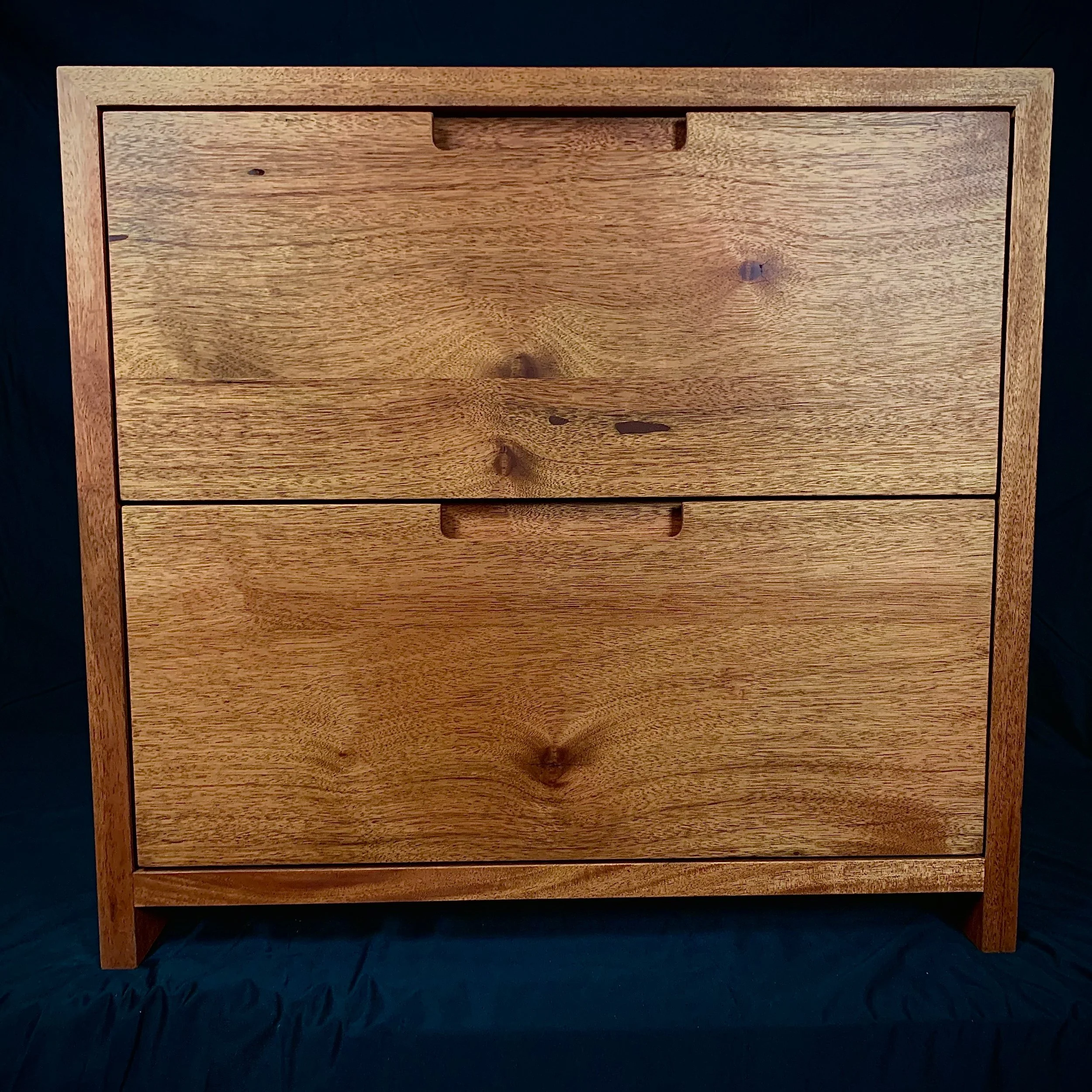 A wooden dresser with three drawers and a natural finish, set against a dark background.