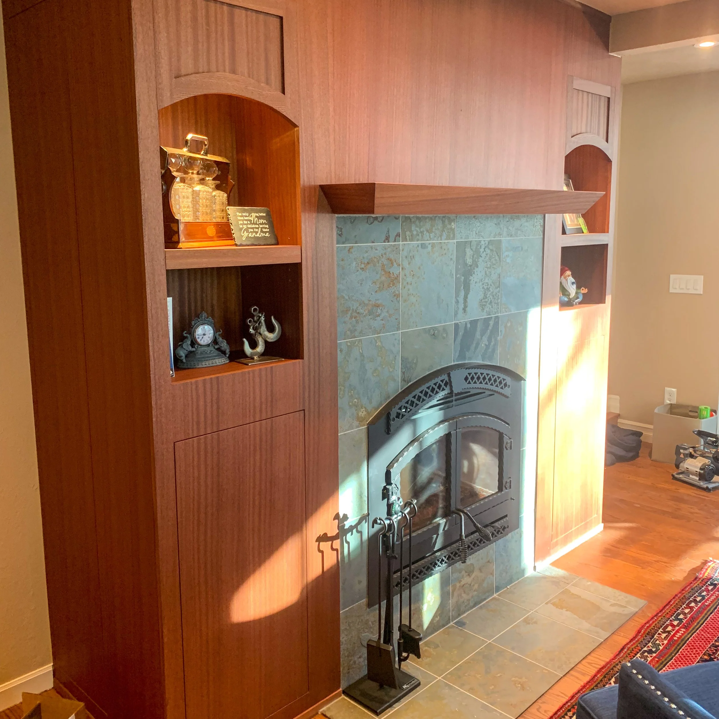 A living room fireplace with a wooden mantle and built-in shelves on each side. The fireplace has a black metal door with decorative ironwork and is surrounded by blue-green tiles. There are small decorative items on the shelves, including a clock, a