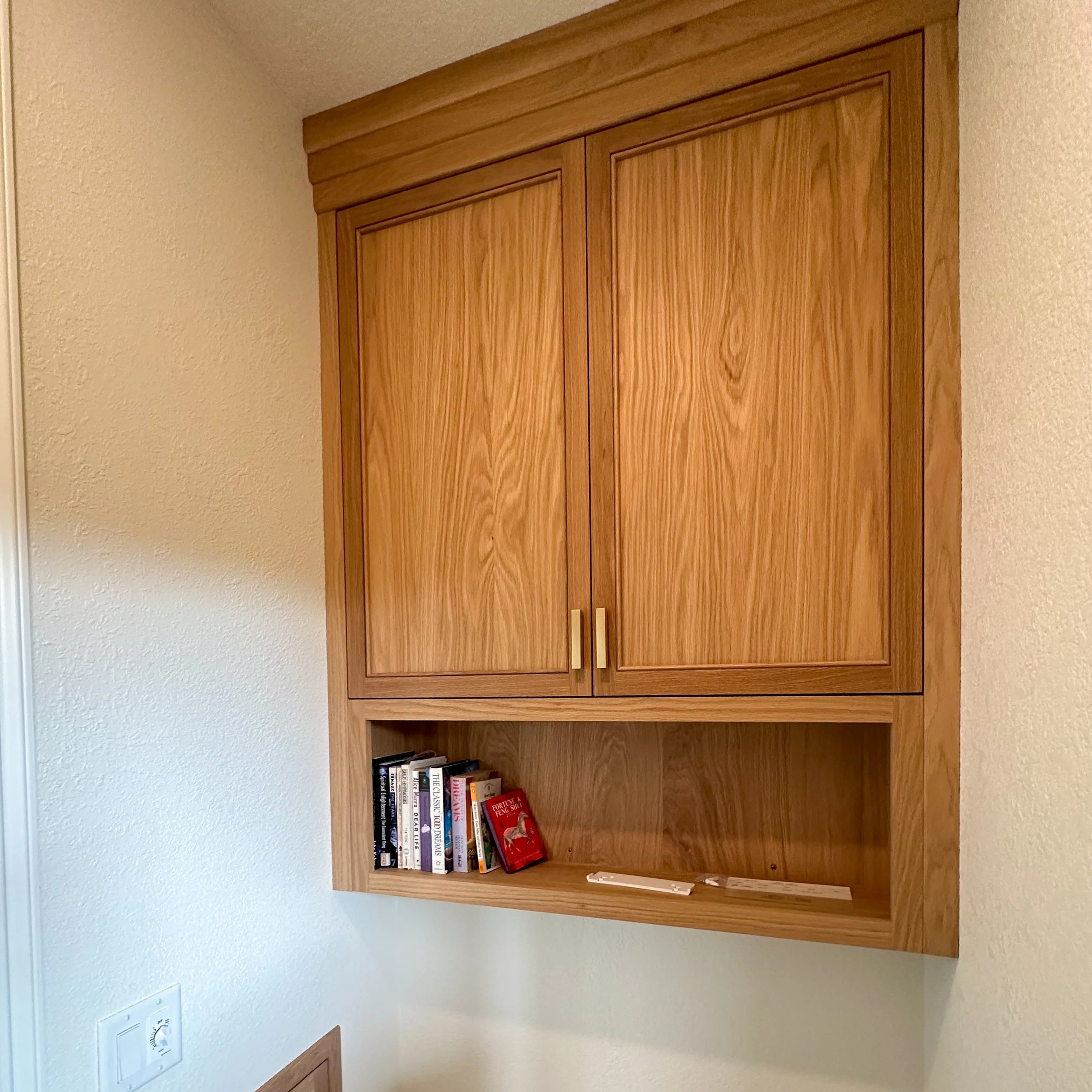 Wooden wall-mounted cabinet with closed upper doors and an open lower shelf holding several books and a small box, against a textured white wall.