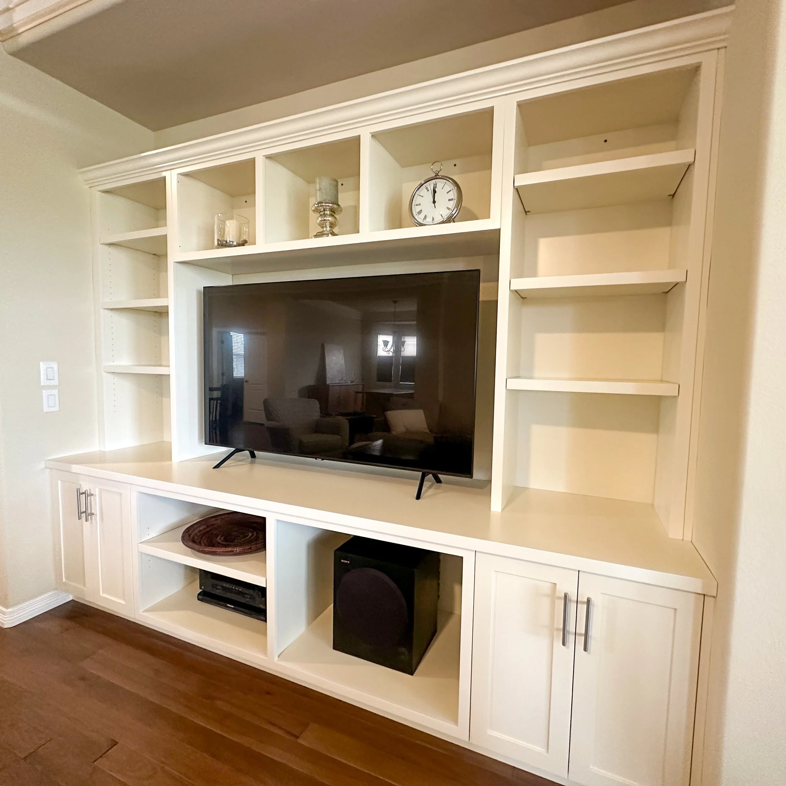 White entertainment center with shelves and cabinets, holding a flat-screen TV, decorative candle holders, a clock, and a speaker on a wooden floor.