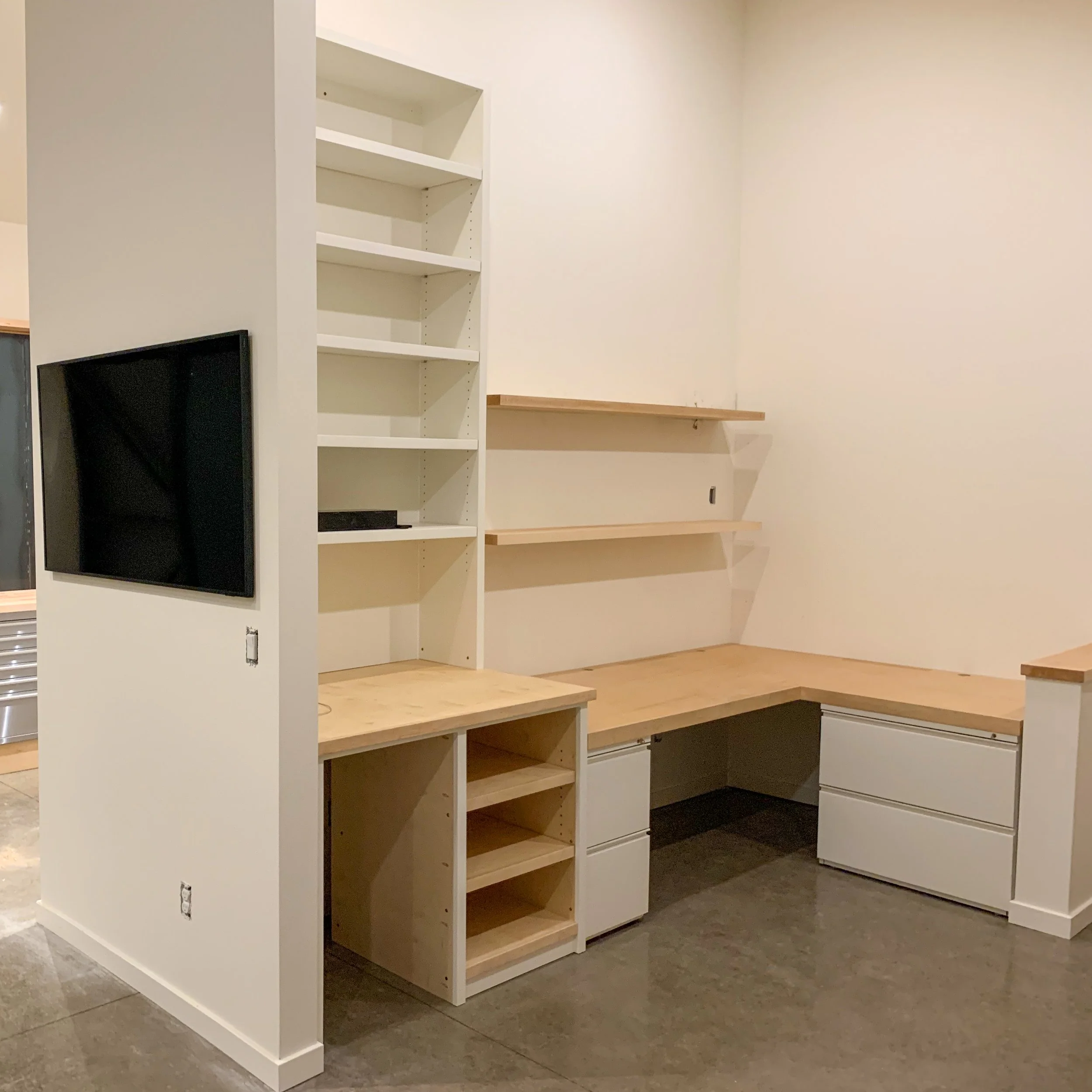 Corner workspace with built-in desk, white shelves, and a mounted TV on the left wall in a room with polished concrete floors.
