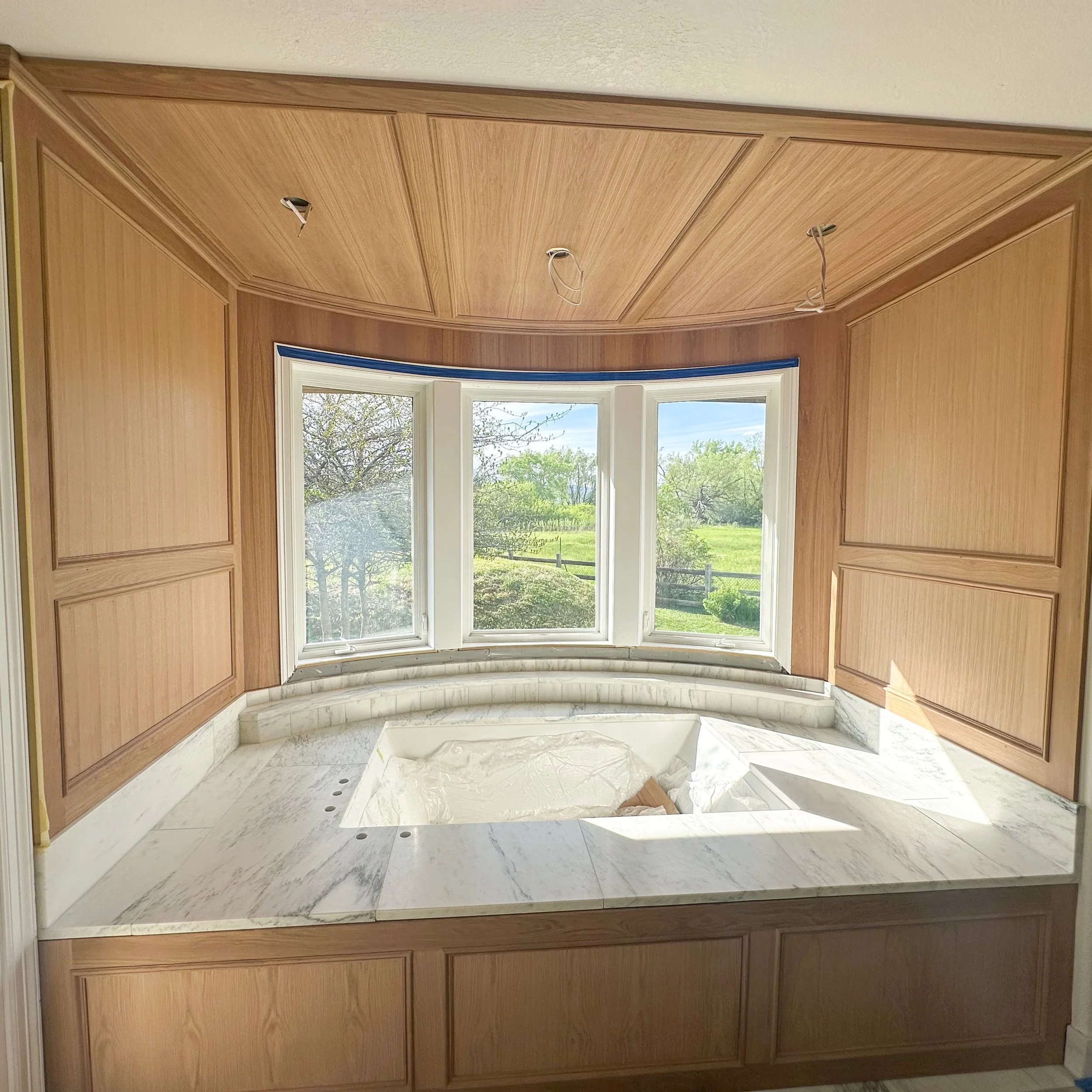 Bathroom with marble countertop and a built-in bathtub below a large triple window, surrounded by wooden wall paneling.
