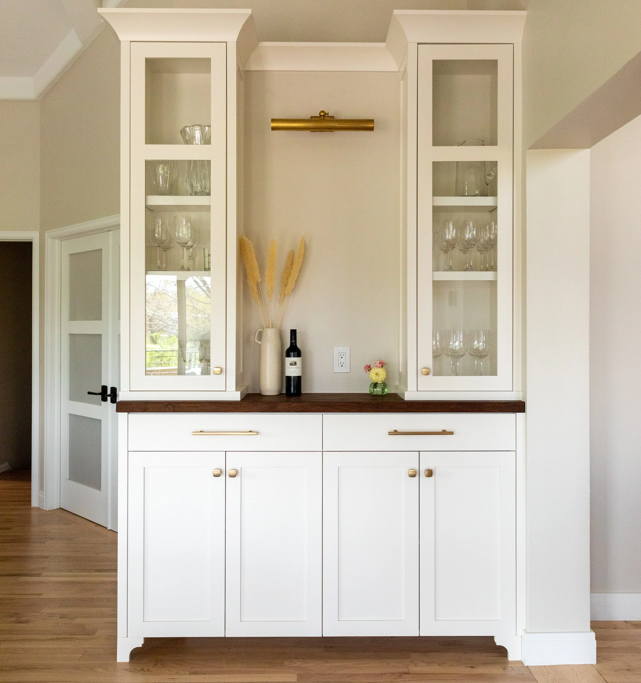 White built-in cabinet with glass doors on the upper section, containing glassware, and closed doors on the lower section, with a wooden countertop. Decor includes dried pampas grass in a vase, a wine bottle, a small flower arrangement, and a wall-mo