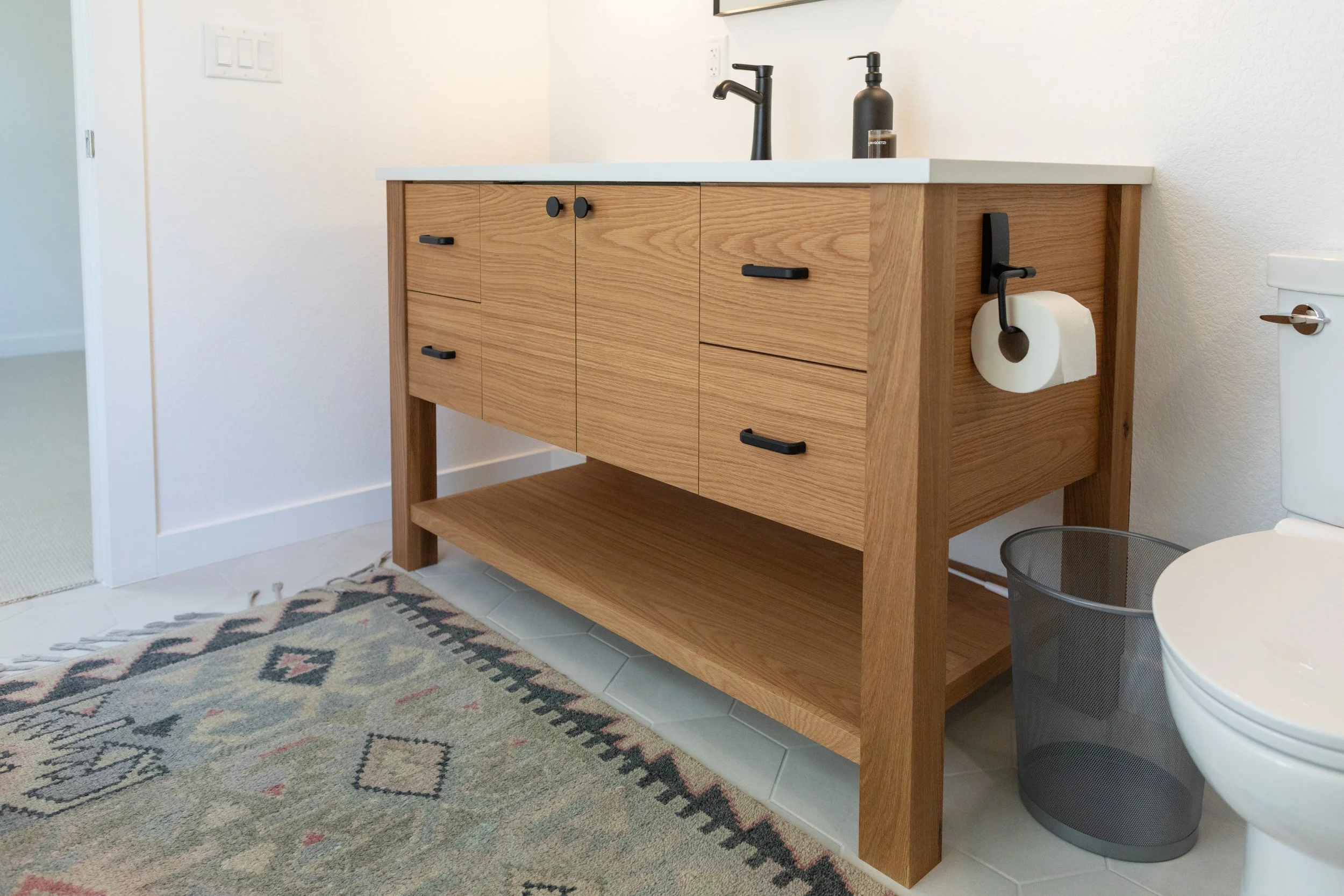 A wooden bathroom vanity with a white countertop, black faucets, a black soap dispenser, and a roll of toilet paper holder on the side, next to a toilet and a black mesh trash bin.