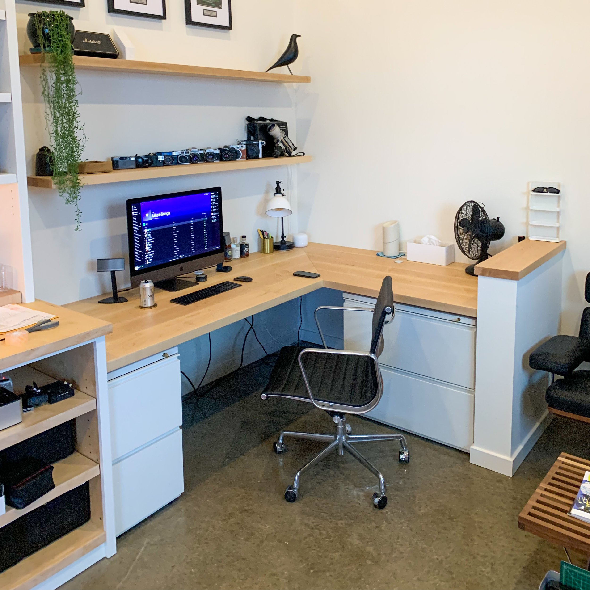 A home office workspace with a wooden L-shaped desk, a desktop computer displaying Spotify, a black office chair, and various items including a lamp, fan, and office supplies. Shelves above hold cameras, decorative objects, and framed pictures.