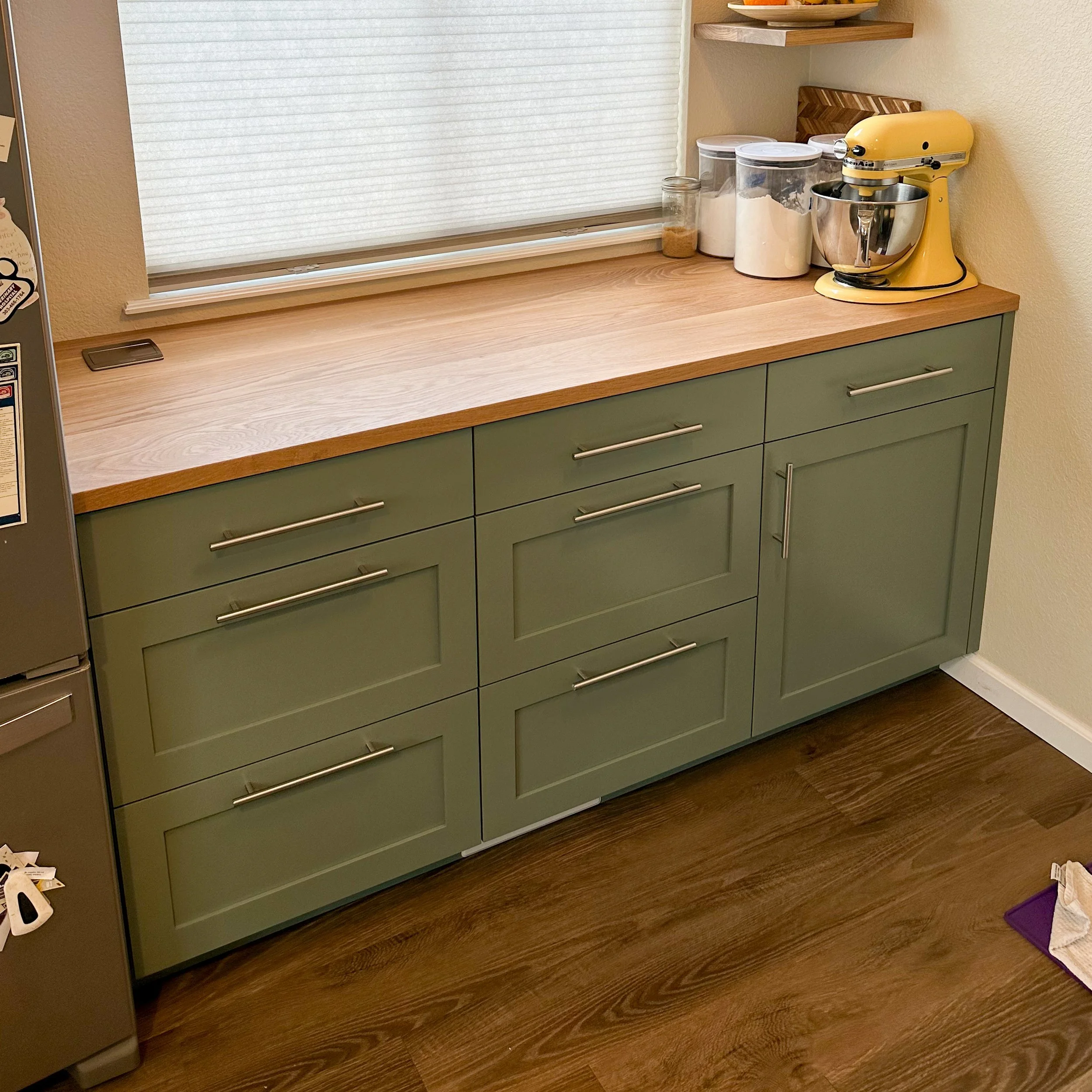 Green kitchen cabinet with silver handles, wooden countertop, yellow stand mixer, jars of flour and sugar, and a window with a white shade