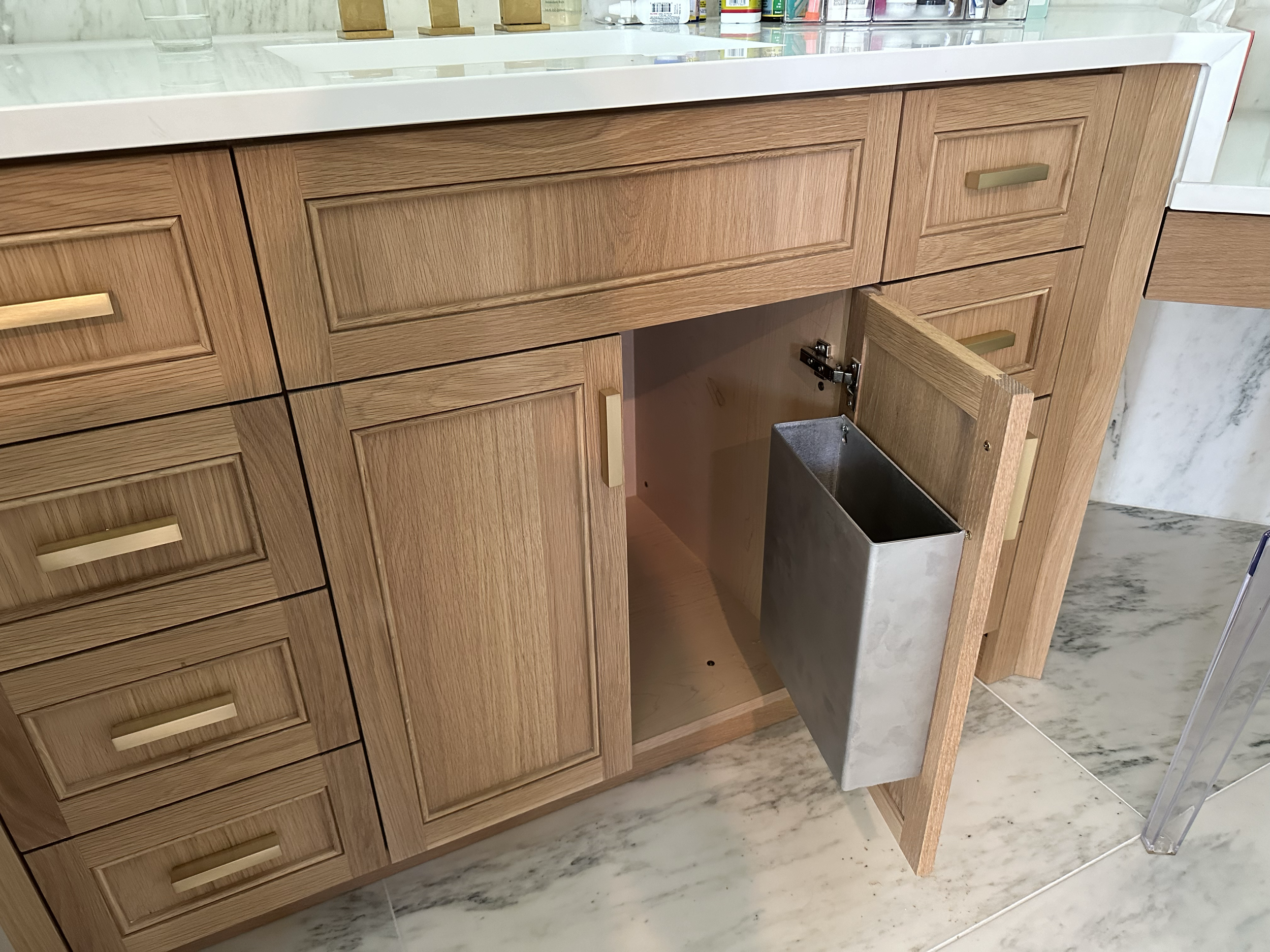 Wooden kitchen cabinet with a drawer partially pulled out, a door open revealing a metal container inside, and a white countertop above.