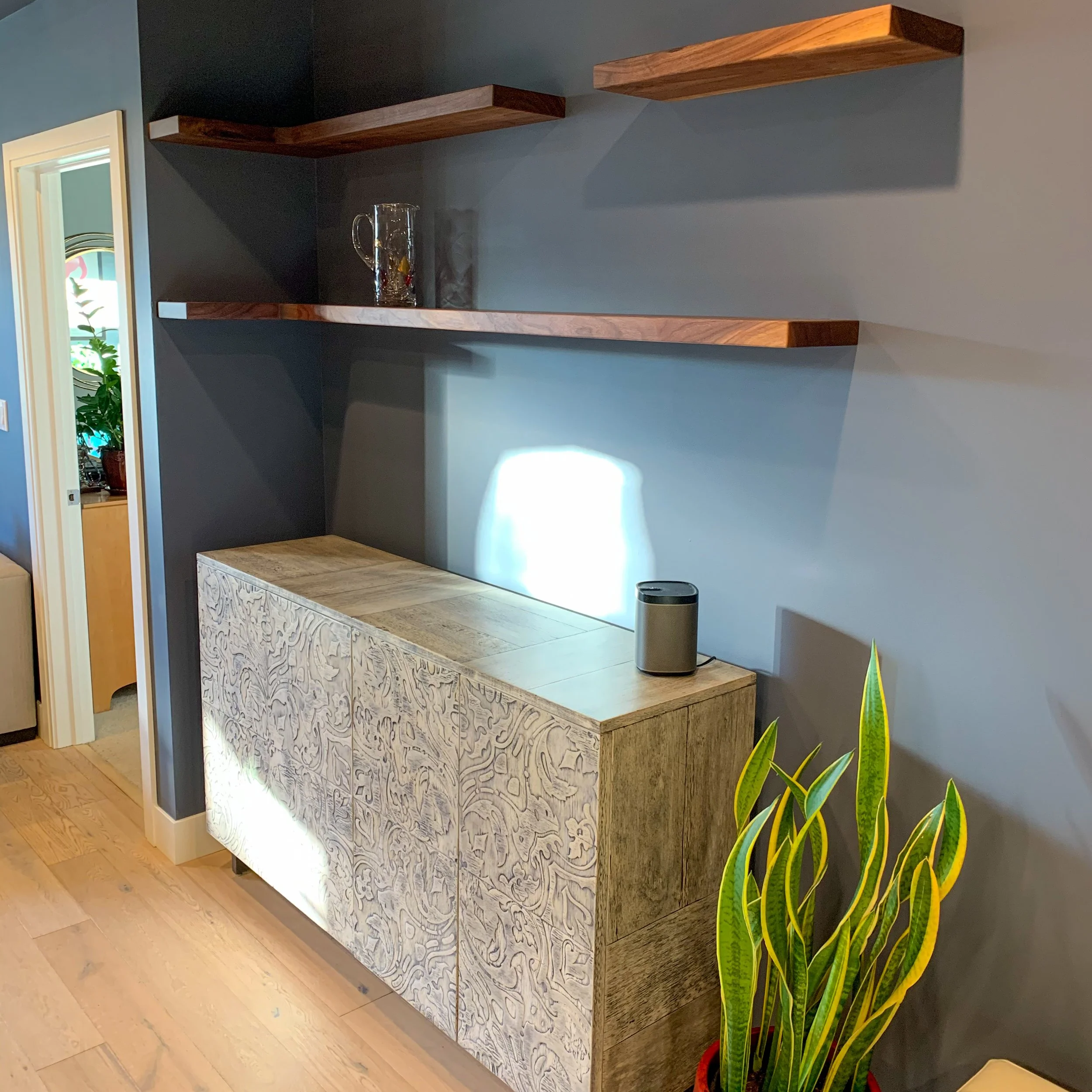 Decorative wooden cabinet with carved floral pattern, topped with a speaker, against a gray wall with two floating wooden shelves, one with a glass decorated jug and the other empty, a plant with long yellow and green leaves in the corner.