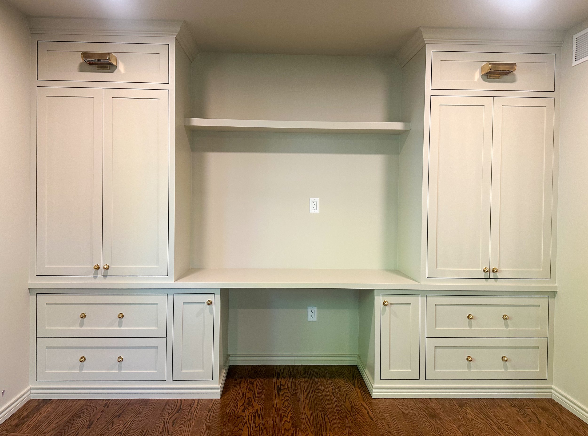 White built-in cabinets with drawers and storage doors, gold knobs, and a central open space with a built-in desk and two electrical outlets. A wood floor is visible.