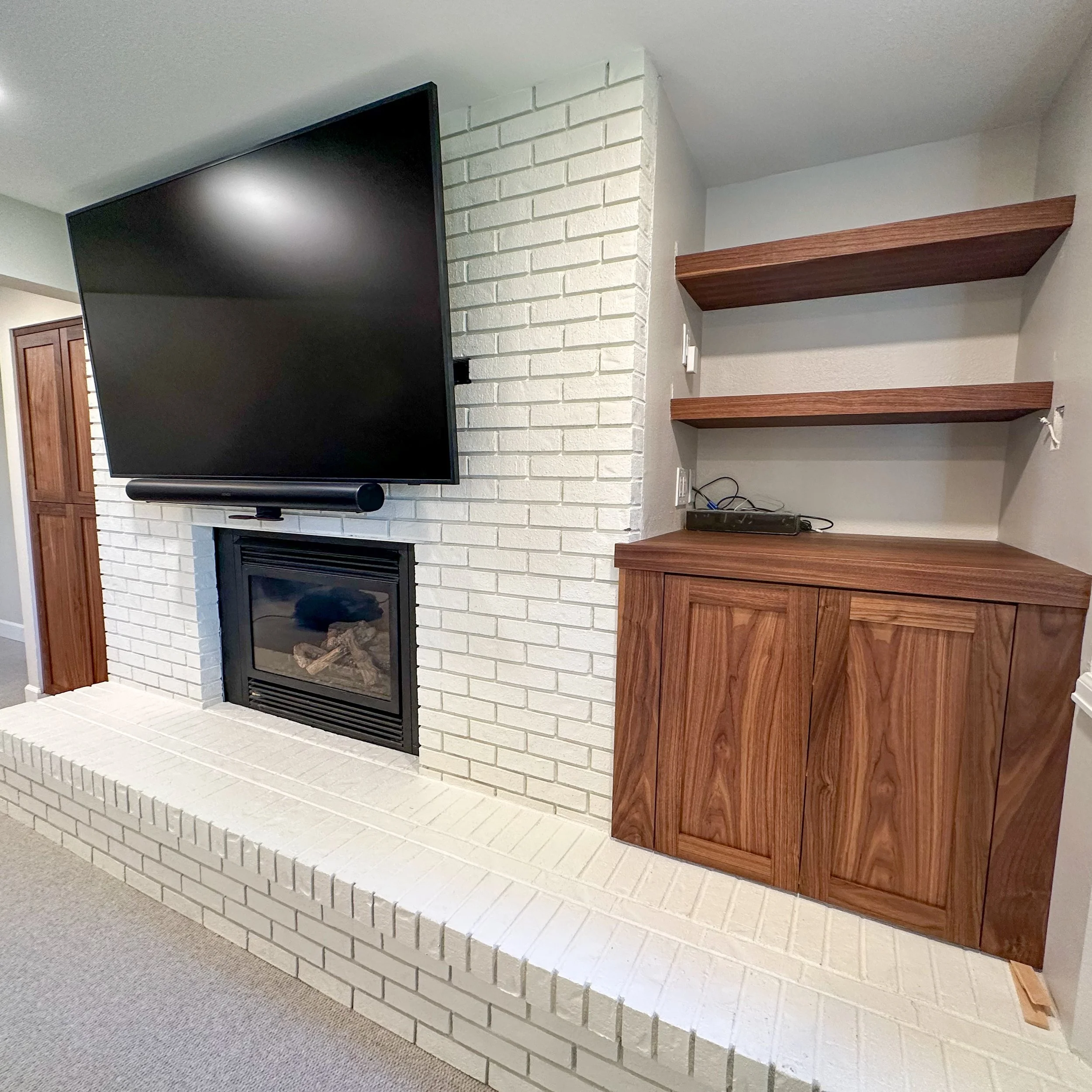Living room with a white brick fireplace, a mounted flat-screen TV, a wooden cabinet, and shelves.