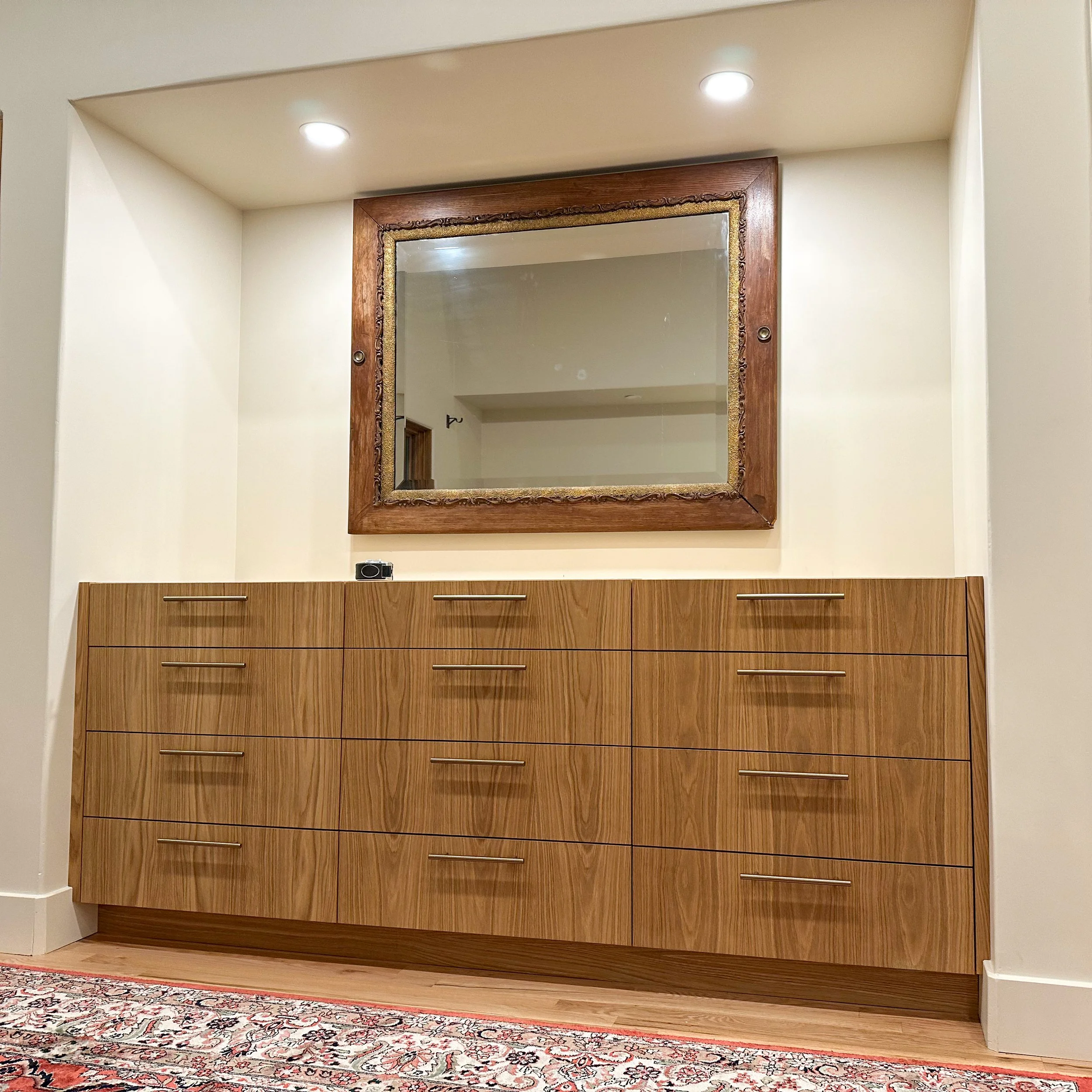 A wooden sideboard with multiple drawers, a large rectangular mirror with a carved wooden frame above it, a small camera or device on the top of the sideboard, and a patterned area rug on the floor.