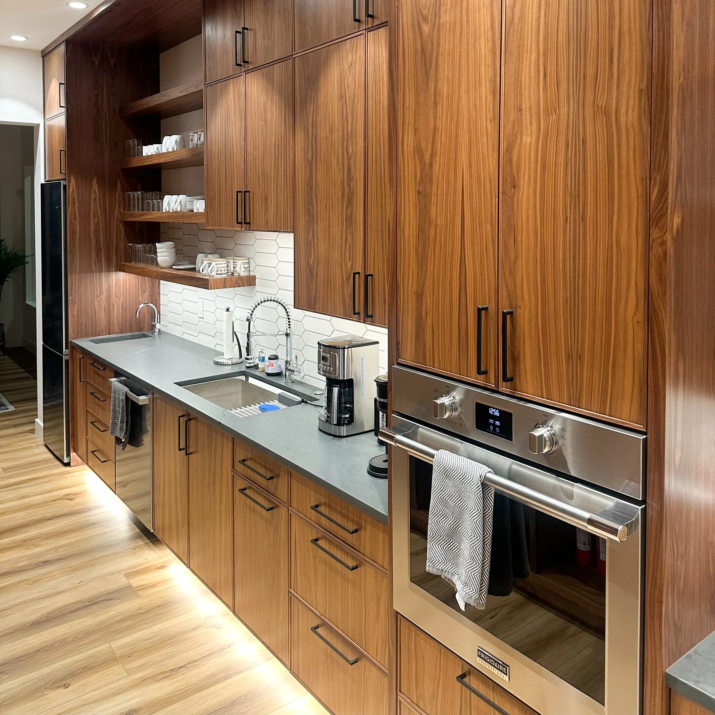 Modern kitchen with wooden cabinets, gray countertop, and stainless steel oven, coffee maker, open shelves with dishes, and a white tile backsplash.