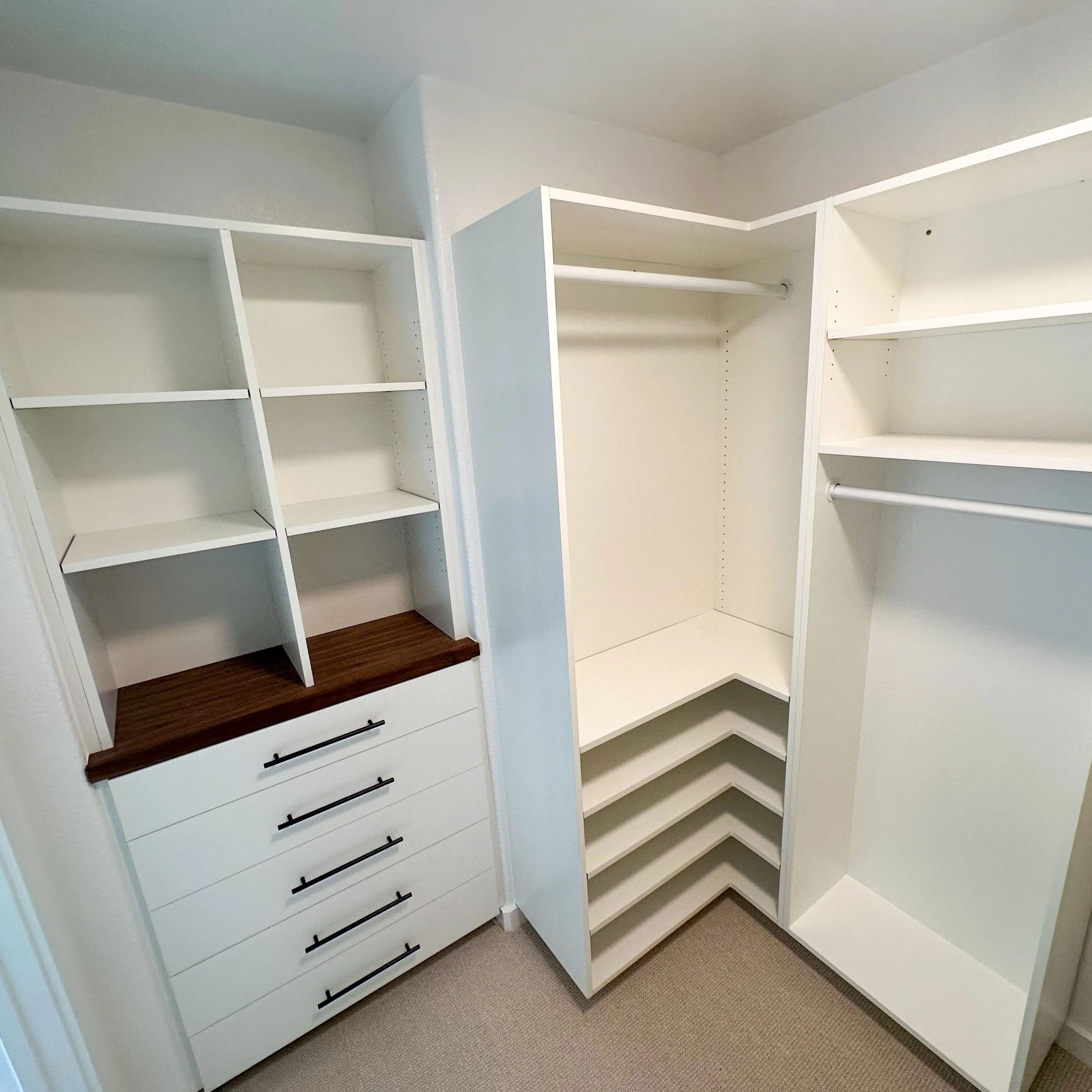 Empty white walk-in closet with shelves, drawers, and hanging rods, with a wood countertop on top of the drawers.