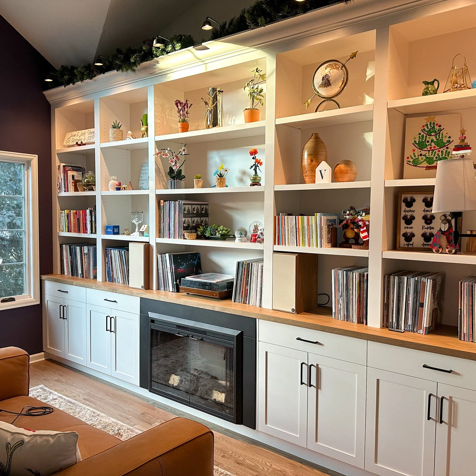 Living room with a large white bookshelf filled with books and decorative items, a window on the left, a brown leather chair, and Christmas decorations along the top of the bookshelf.