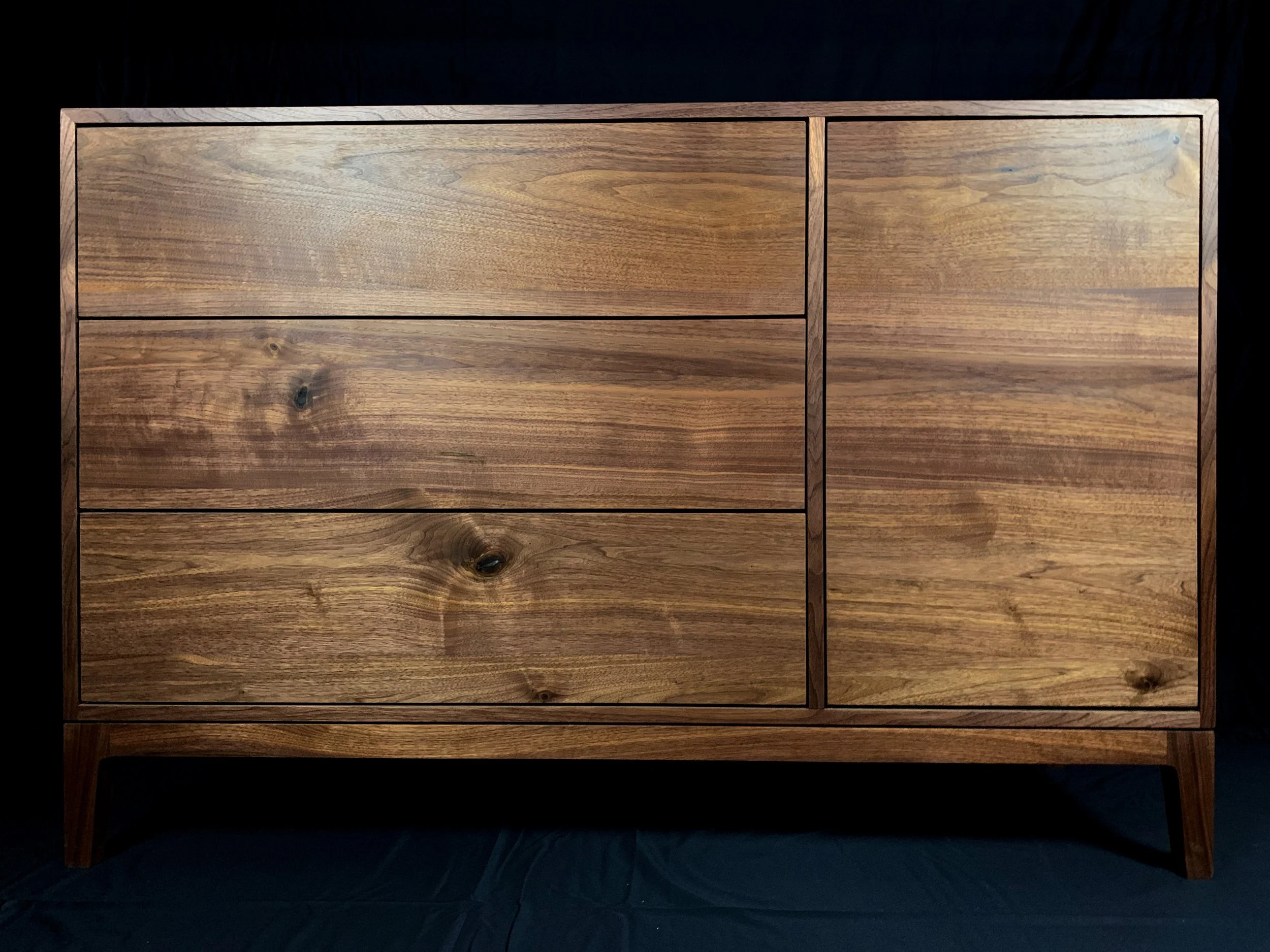 A wooden dresser with four drawers in the left section and a door in the right section, made of natural wood with visible grain patterns, standing on four slightly angled legs.