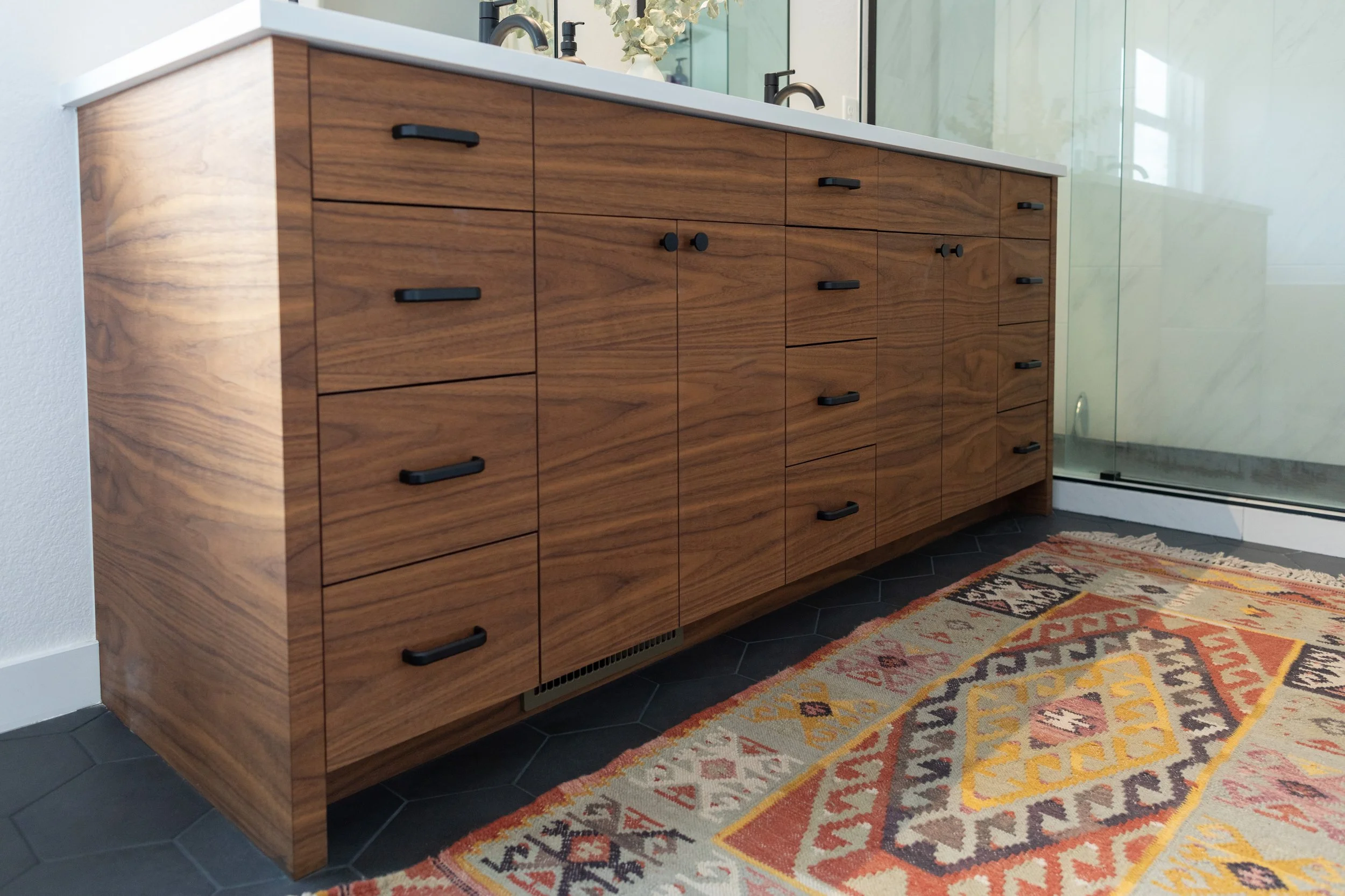 A modern wooden bathroom vanity with black handles situated on a black hexagonal tiled floor, next to a colorful geometric patterned rug, with a white countertop and a large mirror behind.
