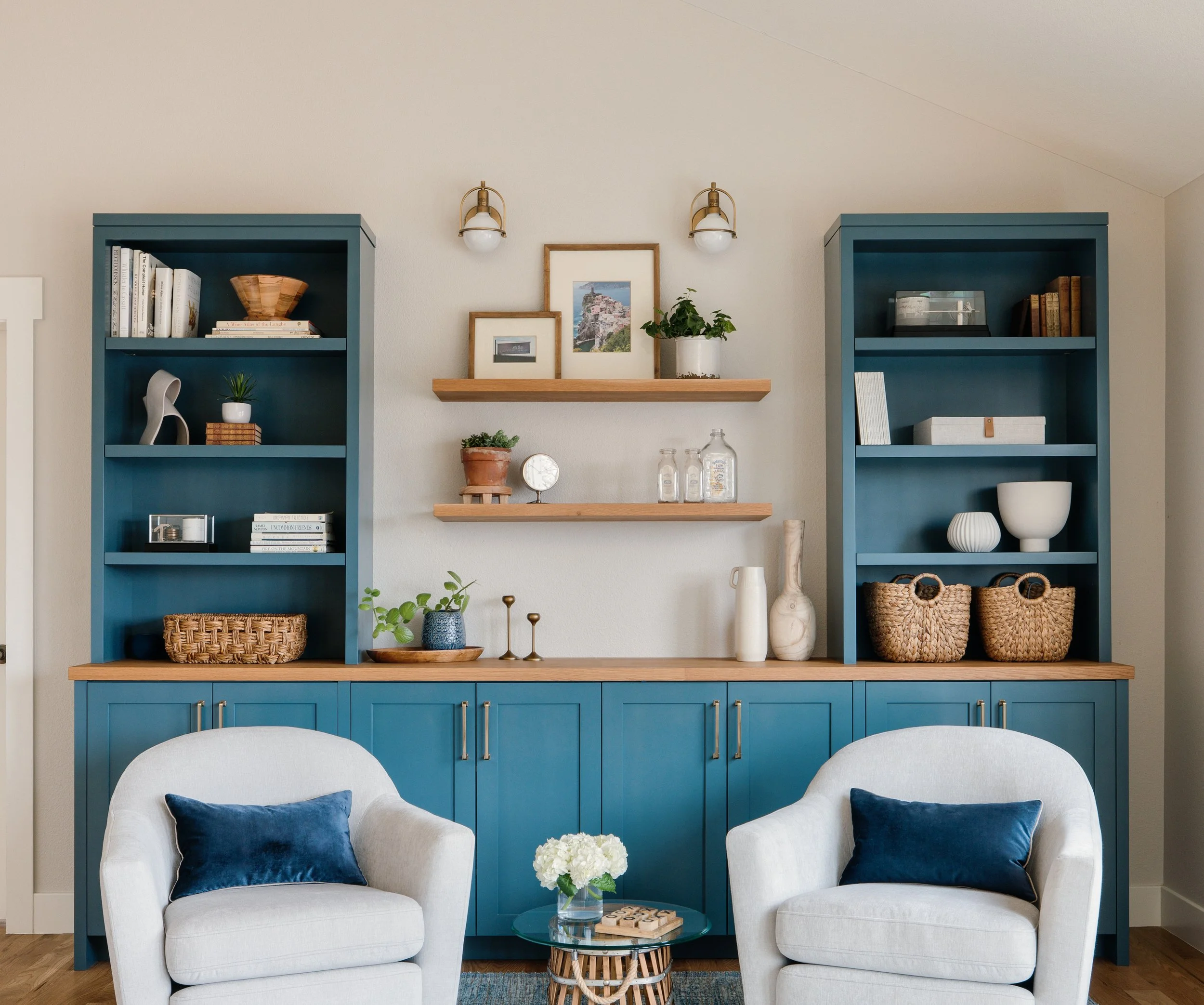 Living room with a built-in blue cabinet, shelves with decor, two white armchairs with navy pillows, and a glass-top side table with flowers.