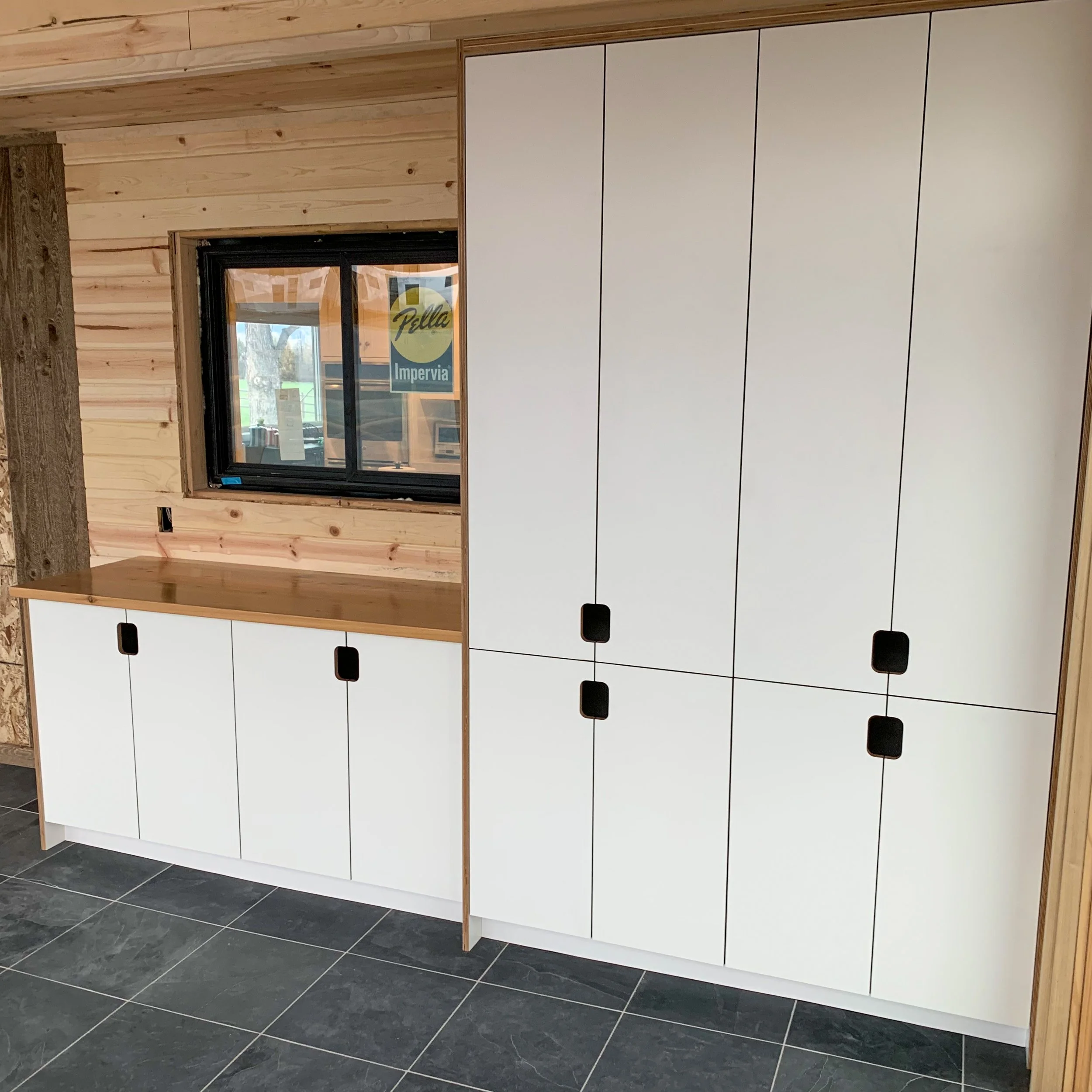 Interior of a room with wooden walls and black tiled floor, featuring a white storage cabinet with black handles and a wooden countertop, along with a window showing an outdoor scene.