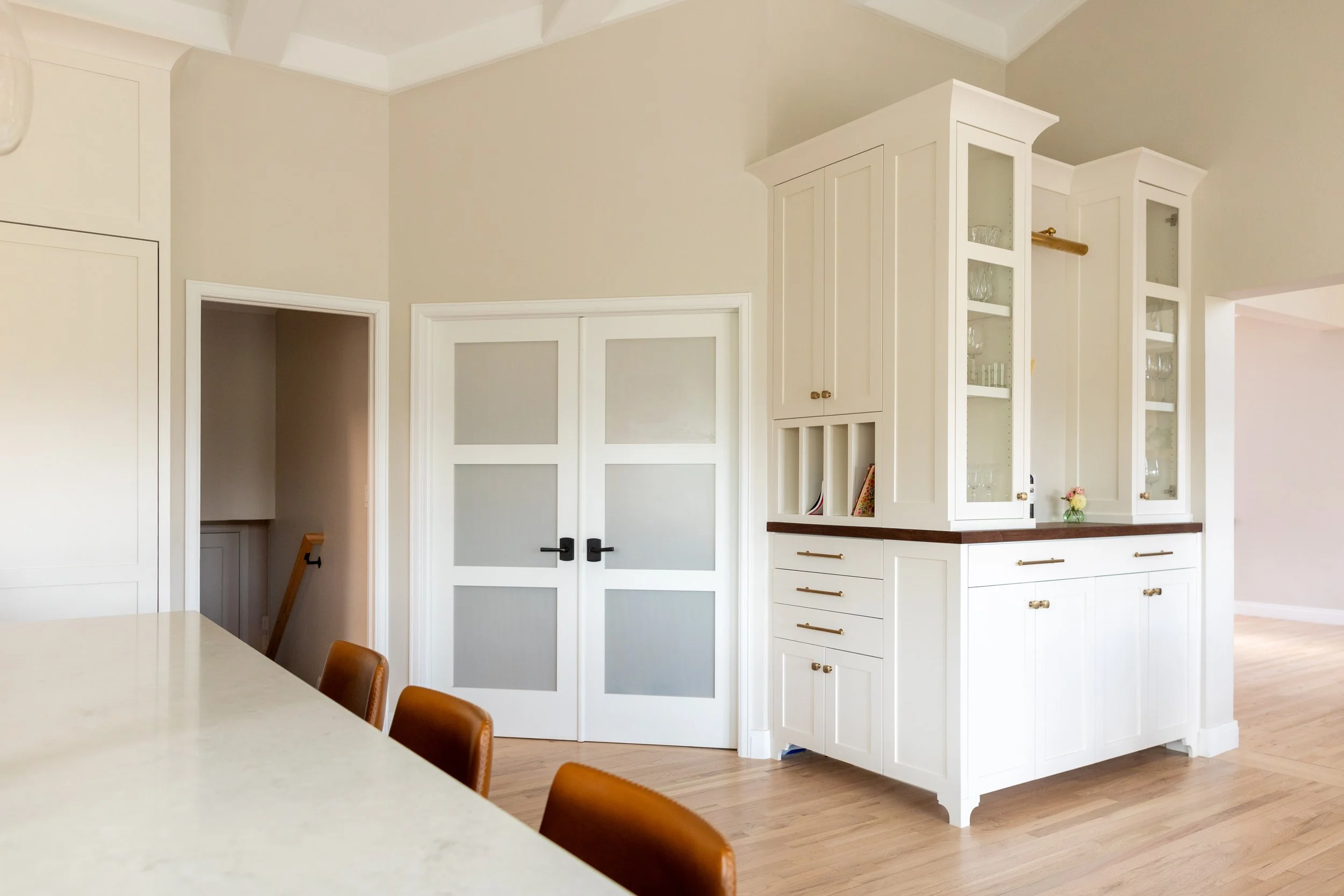 A bright kitchen corner with white cabinets, glass-front display cabinets, and a beige wall. Part of a white countertop and brown dining chairs are visible in the foreground.
