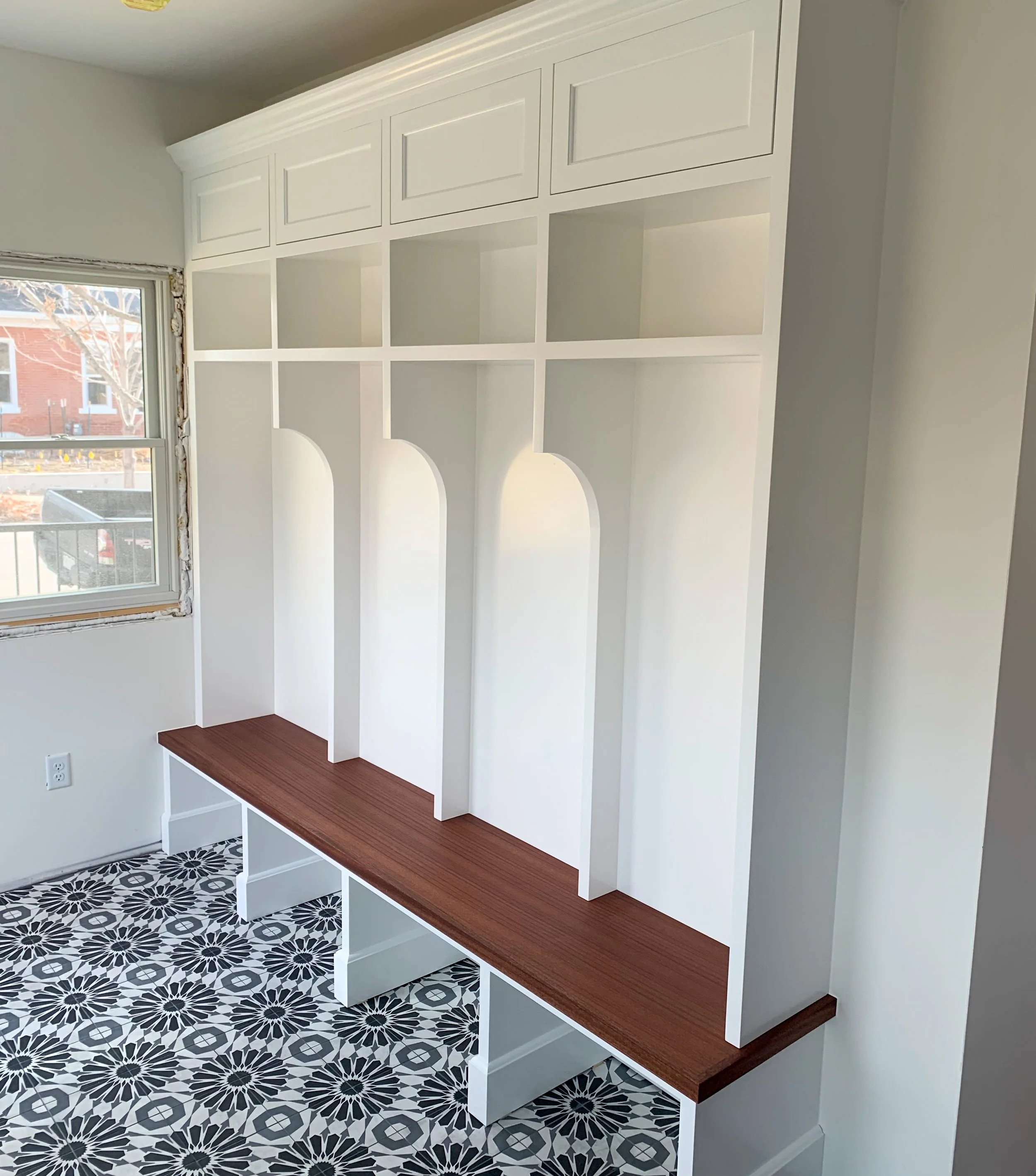 Empty custom built-in white shelving unit with a dark wood bench on a patterned black and white tile floor beside a window in a room.