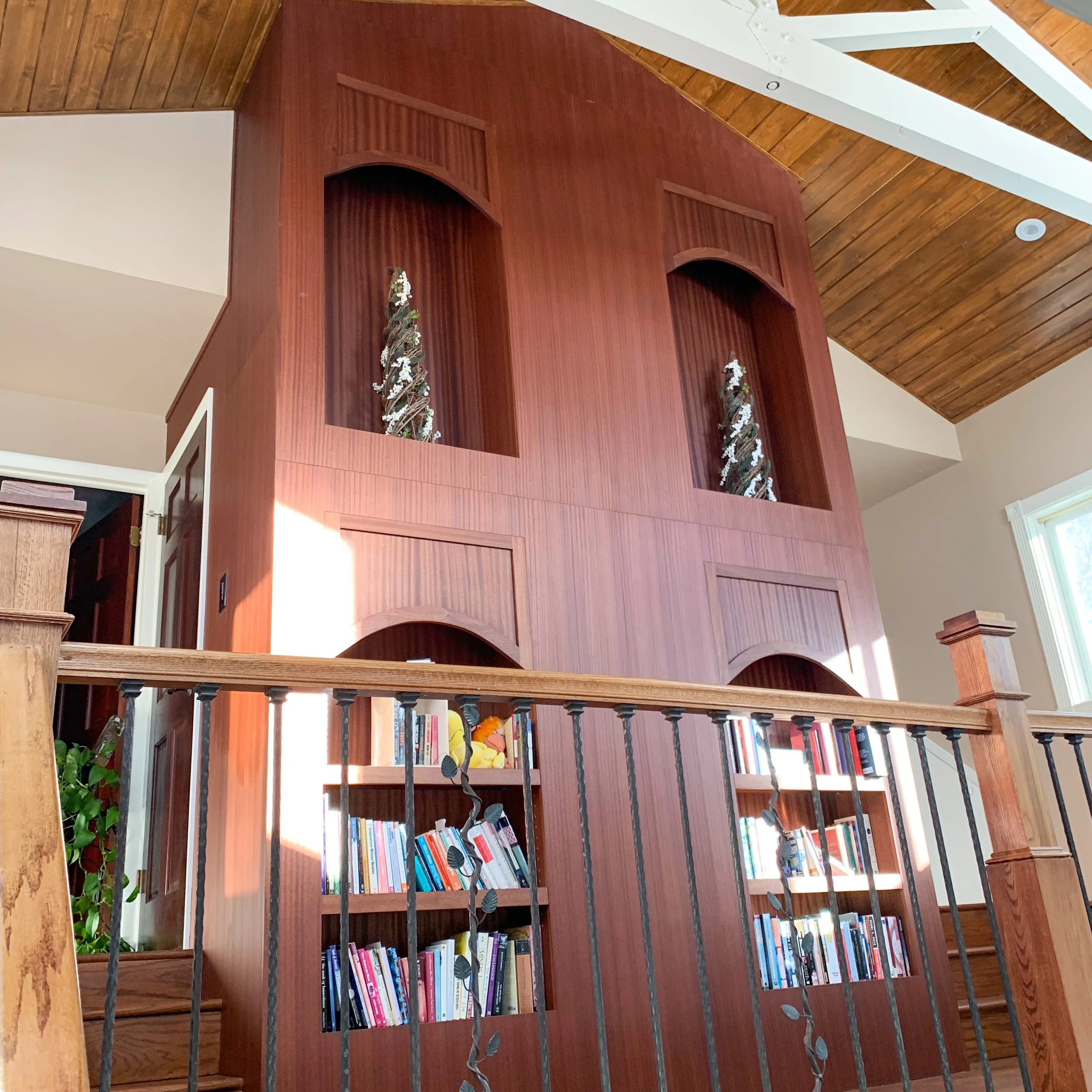 A wooden bookshelf with books and decorations, built into a large wooden wall with arched openings and Christmas trees inside. The room has a high, angled wooden ceiling and a window letting in natural light.