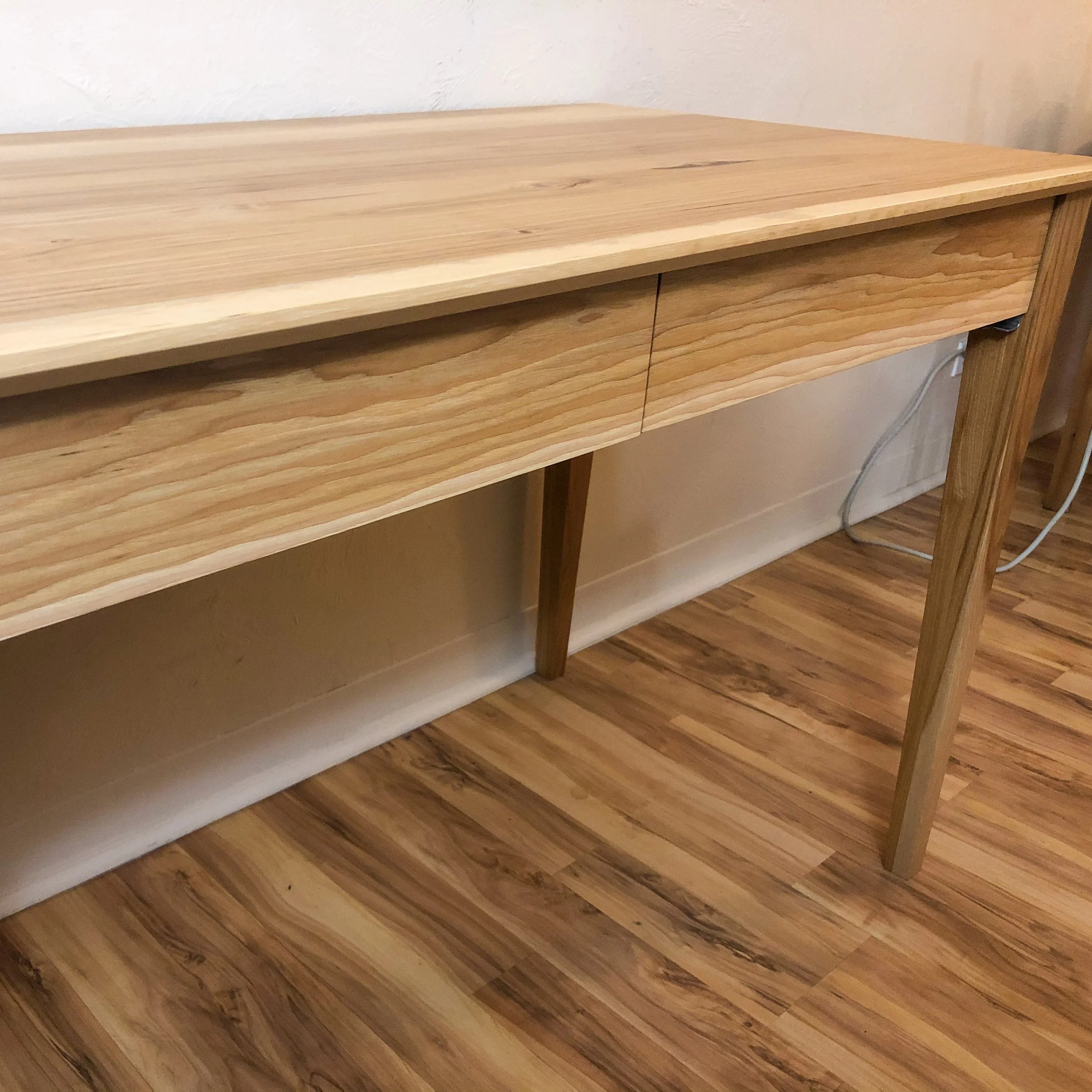 Wooden desk with two drawers, standing on four angled legs, placed against a beige wall with visible electrical cords and outlets.