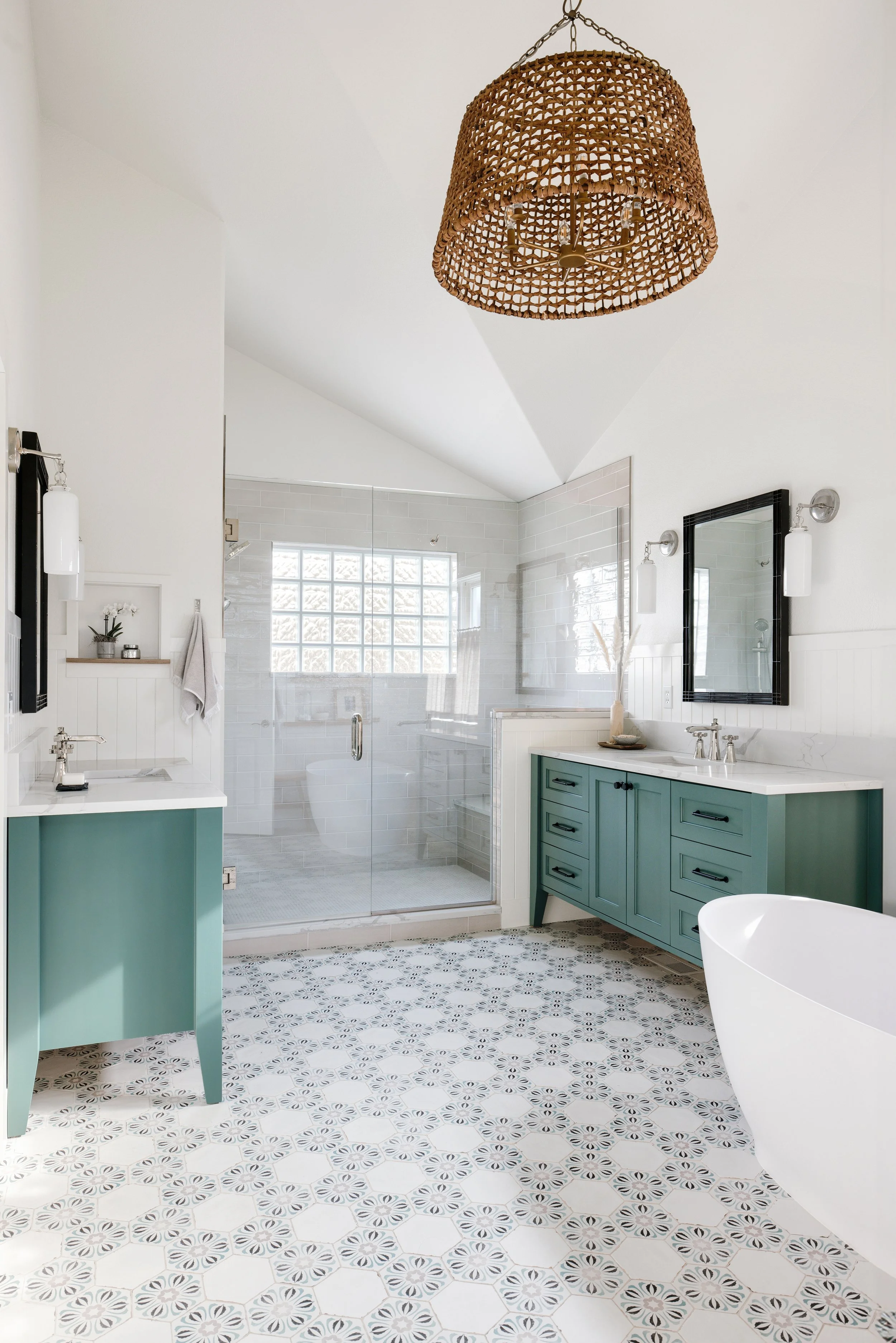 Modern bathroom with two teal vanities, a glass-enclosed shower, a freestanding bathtub, patterned tile flooring, a woven pendant light, and white walls.