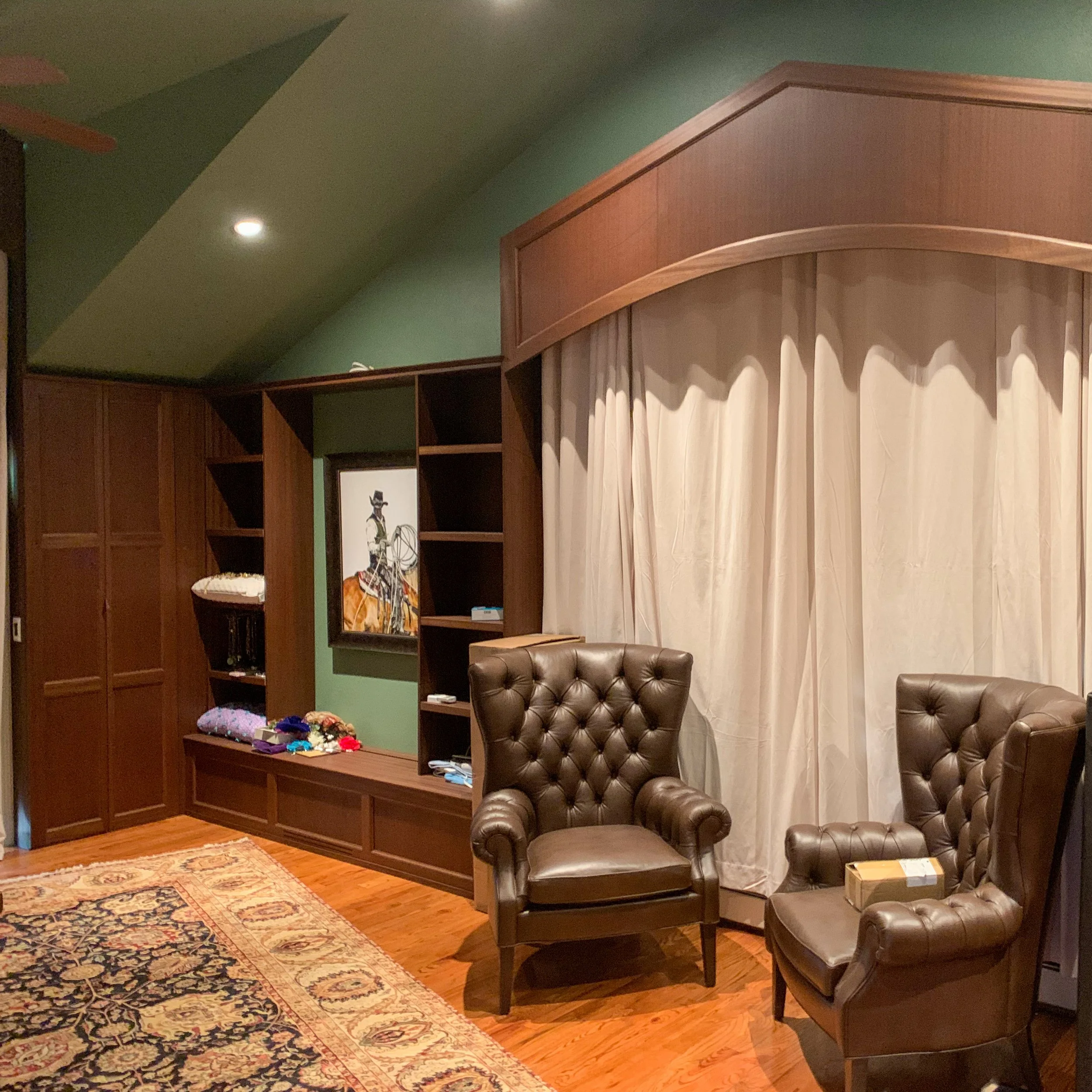 A cozy room with green walls, wooden flooring, and leather armchairs. There is a built-in wooden shelving unit with a photo or painting of a person in formal attire on the wall and various small items on the shelves. A large curtain covers the window