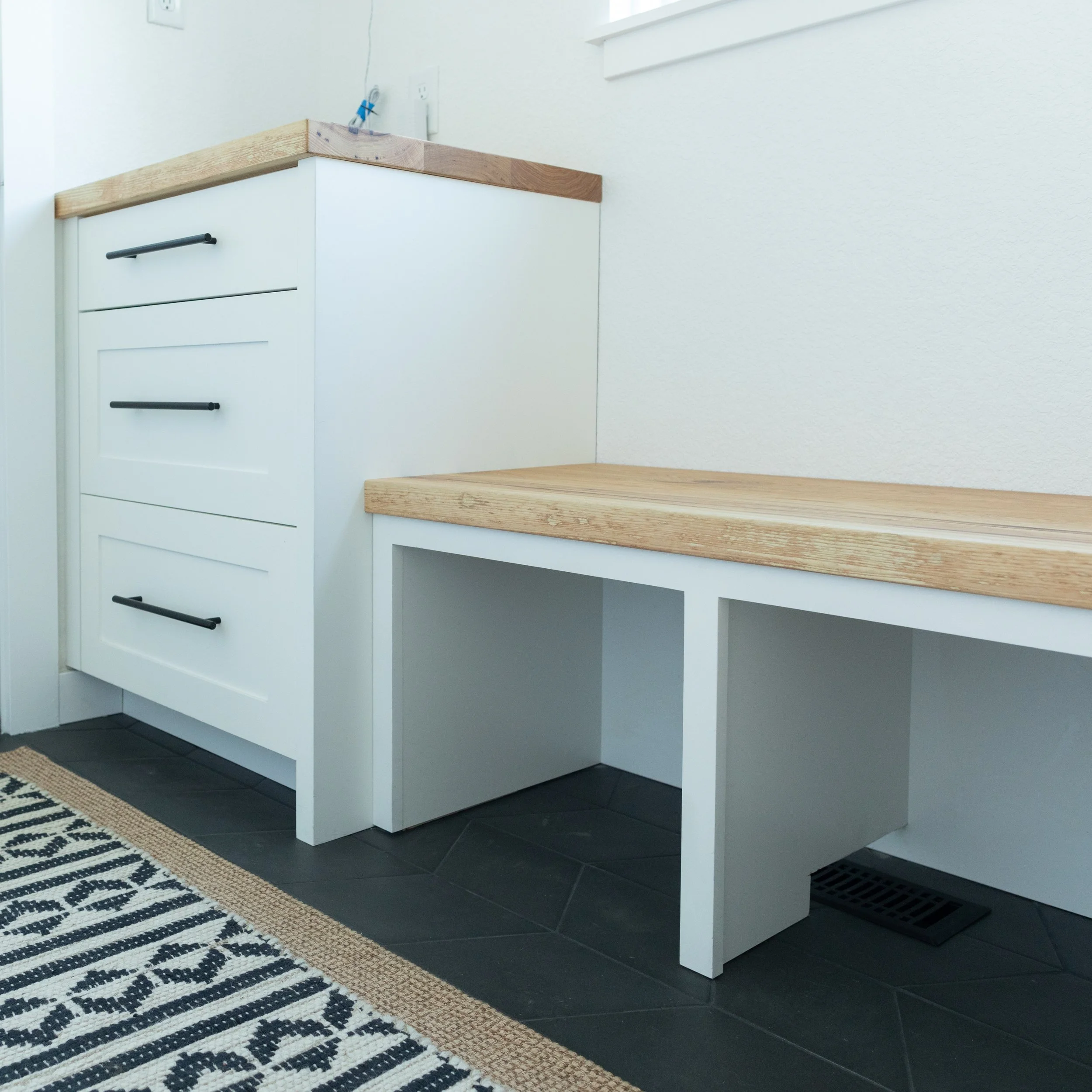White bench with a wooden top next to a white cabinet with three drawers and black handles, located in a room with black tiled flooring and a patterned black and white rug.