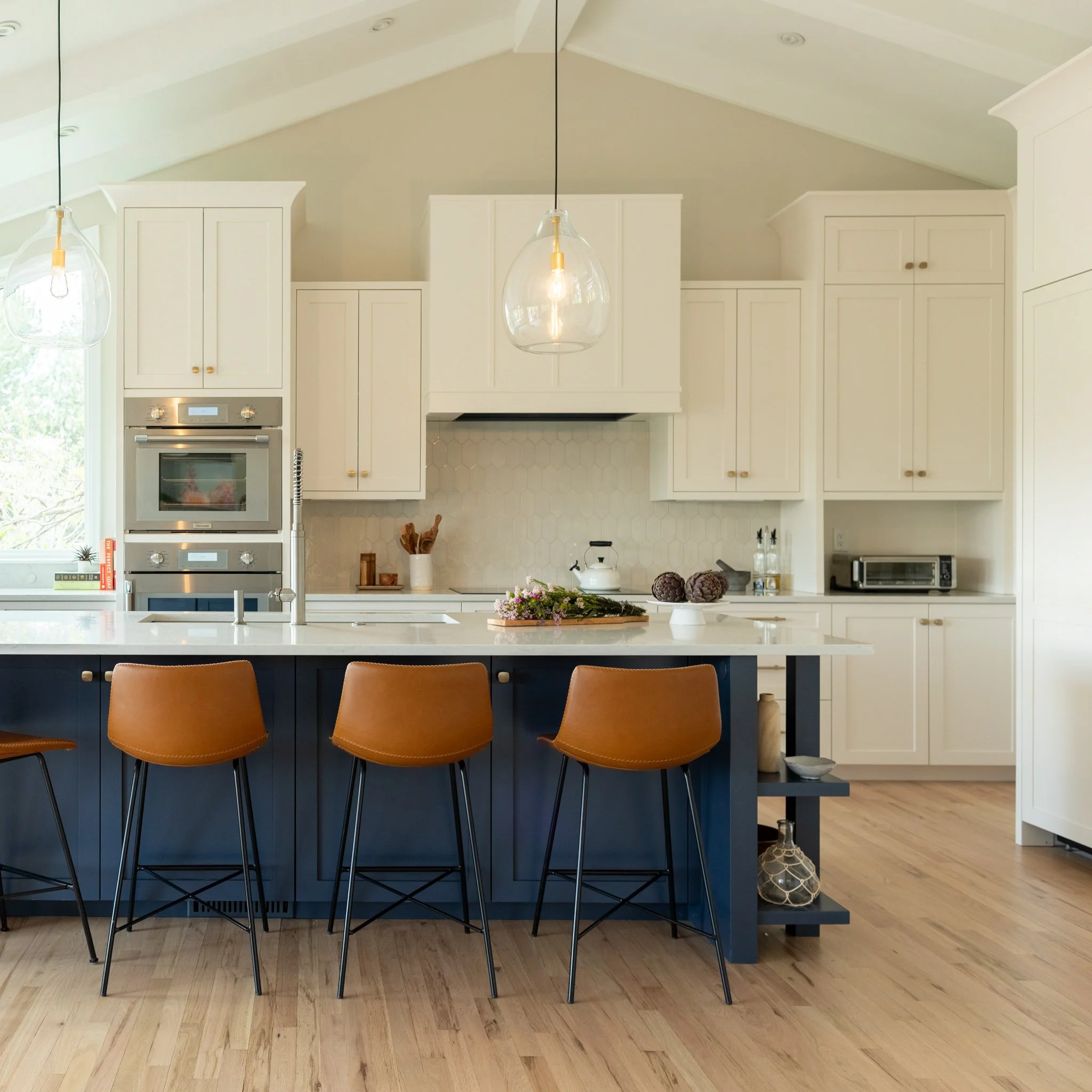 Modern kitchen with white cabinets, a navy blue island with three brown leather barstools, pendant lighting, and light wood flooring.