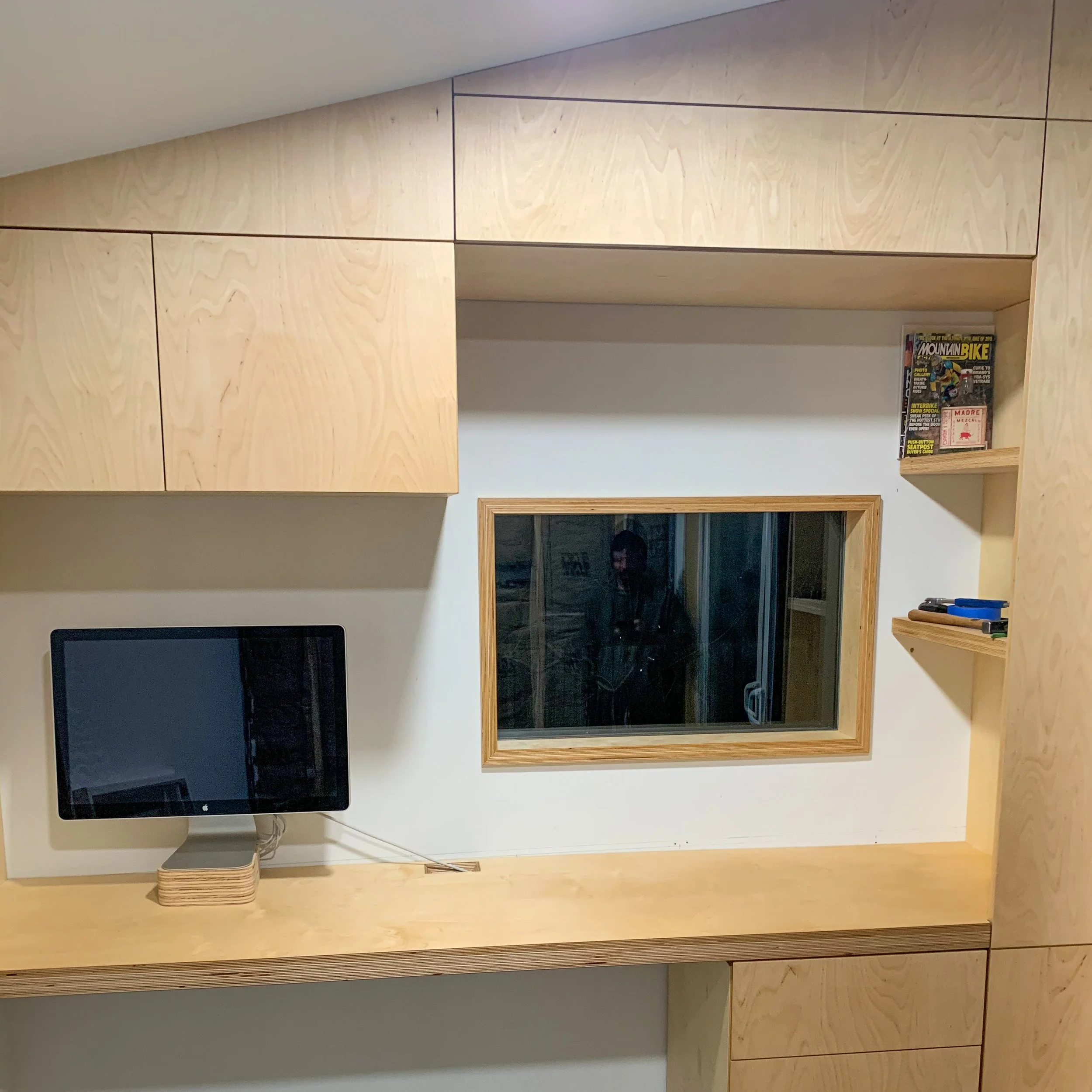 Interior view of a room with light wooden shelves, cabinets, and a countertop. A small computer monitor is on the desk, and there is a window with a person's reflection visible in the glass.