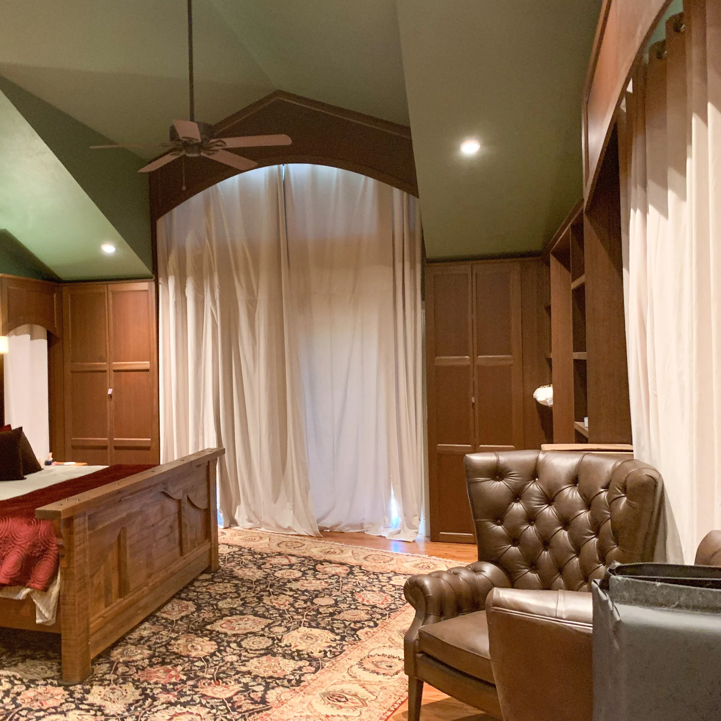 A bedroom with a wooden bed, leather armchair, and built-in wooden shelves and cabinets, white curtains covering a large window, and a patterned area rug on the floor.