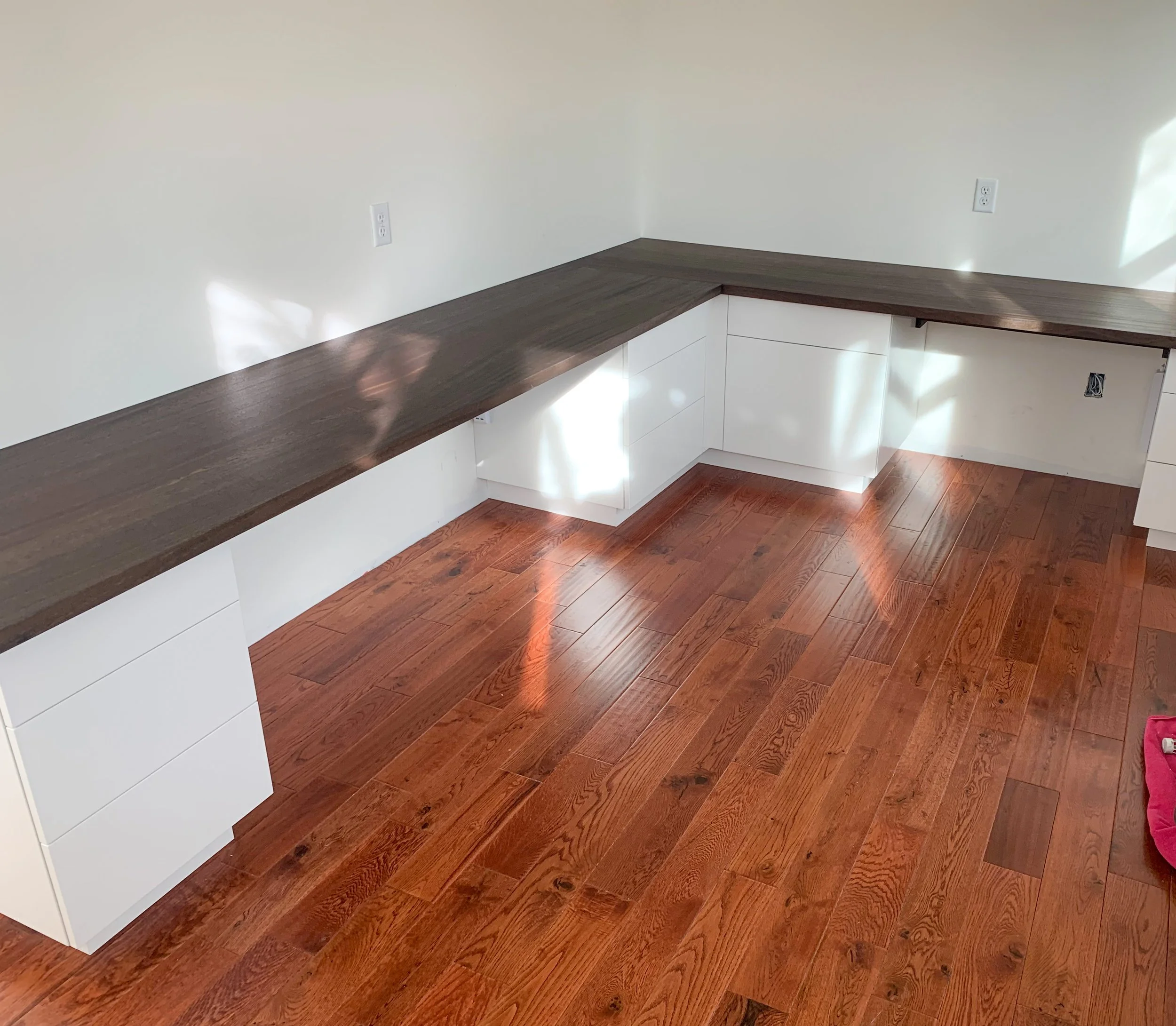 A corner of a room with a wooden floor, white walls, and a built-in L-shaped desk with a dark wooden surface and white cabinets underneath.