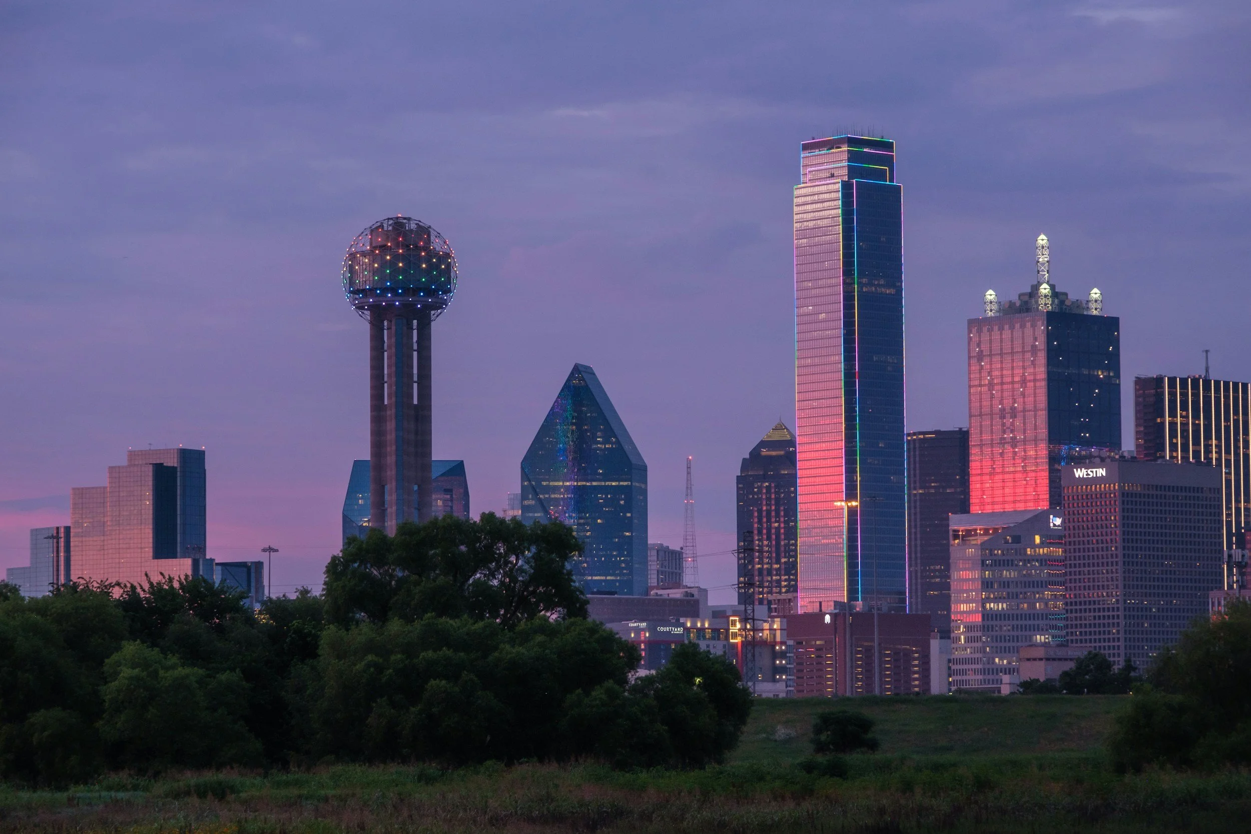 Skyline of Dallas, Texas at dusk, with illuminated skyscrapers including the Reunion Tower on the left and the Bank of America Plaza on the right, seen from a distance with trees in the foreground.