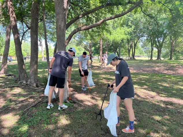 People cleaning up litter in a park with trees and grass, some wearing hats and casual clothes.