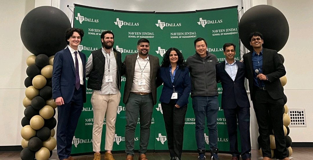 Seven people in formal attire pose at a UT Dallas Naveen Jindal School of Management event in front of a green backdrop with balloon columns.
