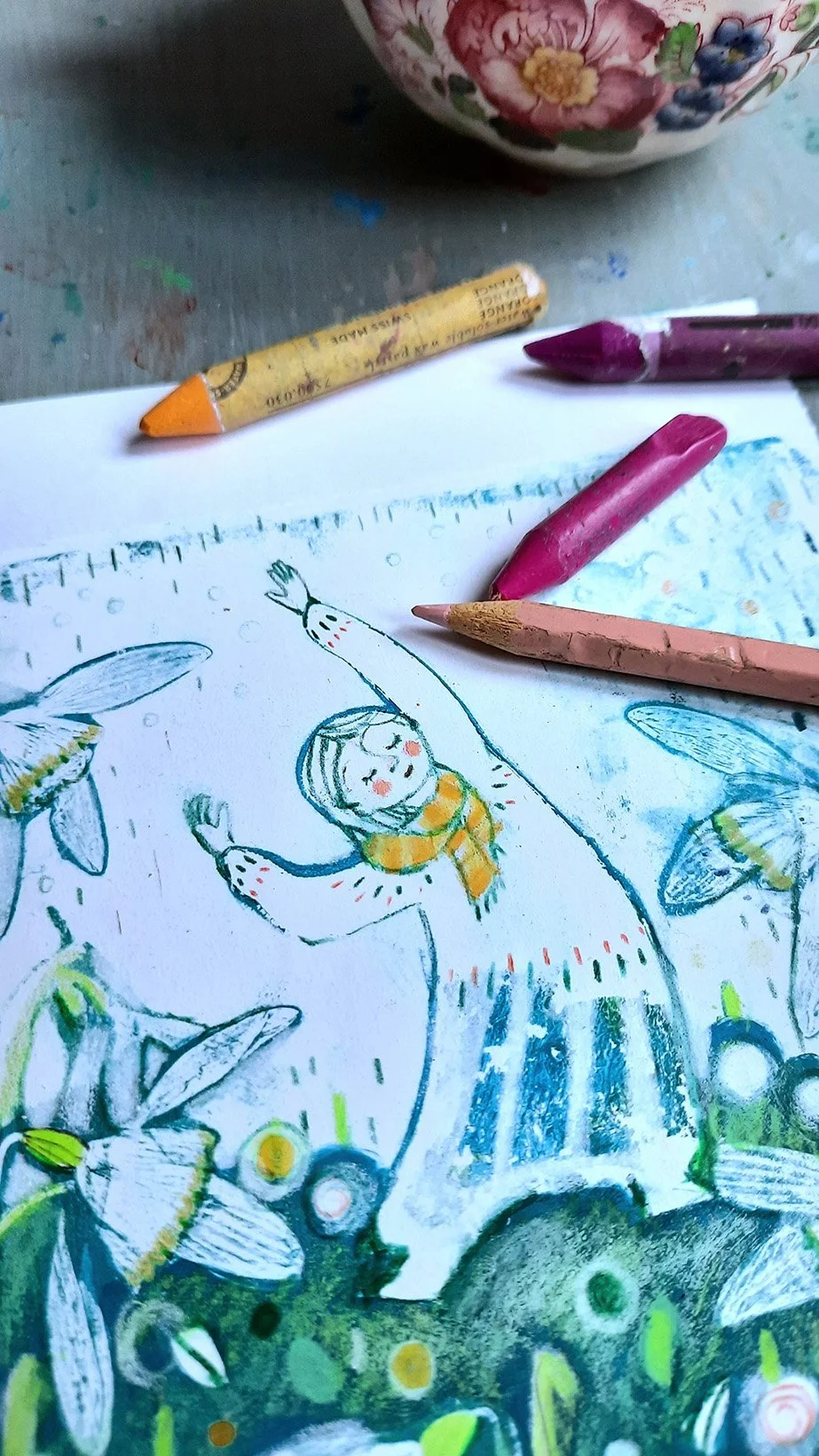Drawing of a girl with a yellow scarf surrounded by bees, with colored crayons scattered around, on a table.