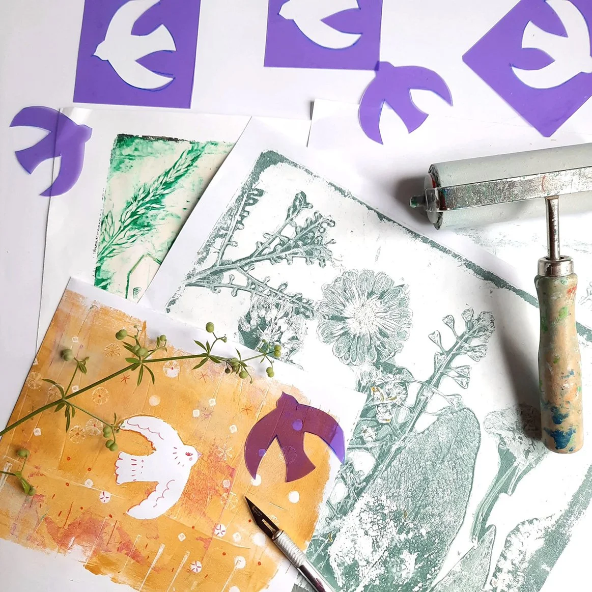 A printing workspace with botanical printouts, purple decorative cutouts, a printing roller, and a pen. The workspace has botanical illustrations of plants and flowers, some in green and black and white, and a colorful print featuring a bird and flow