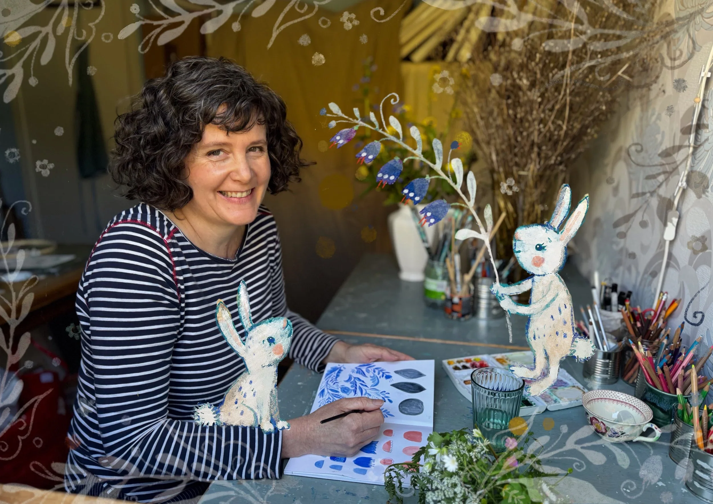 Photo of the artist in her studio painting leaf motives with one small white bunny on her arm and another bunny opposite her modelling a leafy twig with blue bells on it.