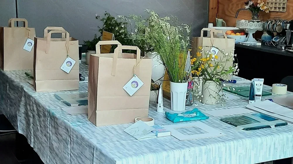 Table set with art materials, botanicals in vases and brown paper bags, ready for a botanical printing workshop.