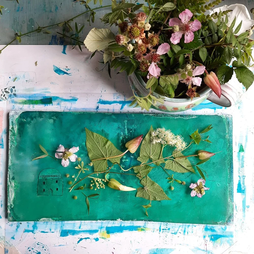 Artistic arrangement of various flowers and greenery on a teal surface, with a bouquet of mixed flowers in a mug on a white, paint-spattered table.