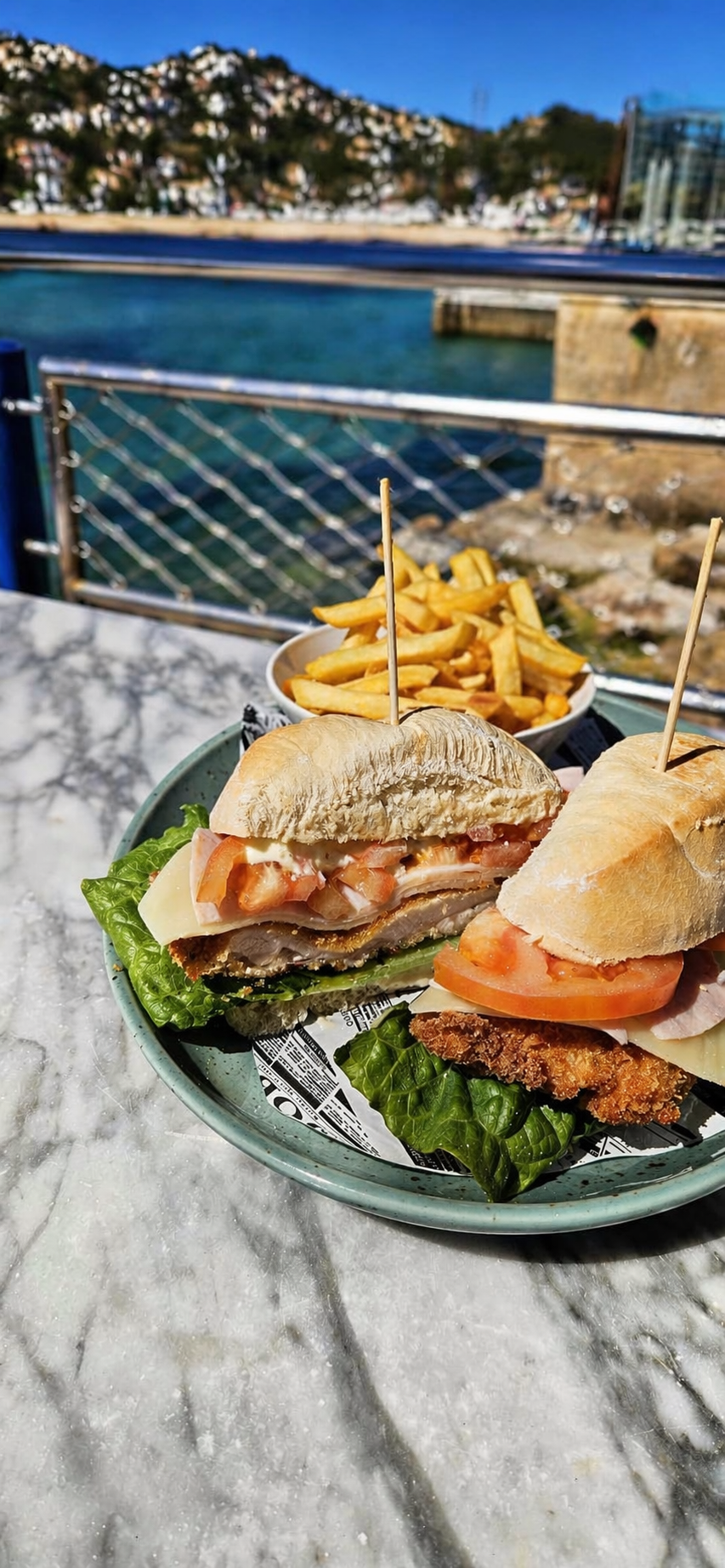 Sandwiches con papas fritas en un restaurante con vista al mar y una colina con casas en el fondo.