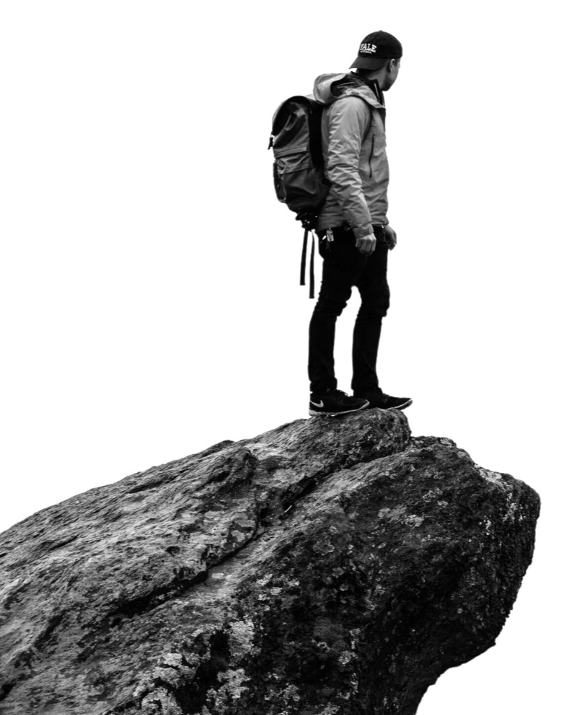 A man with a backpack standing on a large rock or cliff edge, gazing towards the horizon, dressed in outdoor gear, black and white photo.