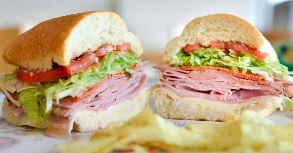 Sandwich with ham, lettuce, tomato, and cheese on a sub roll, served with potato chips.