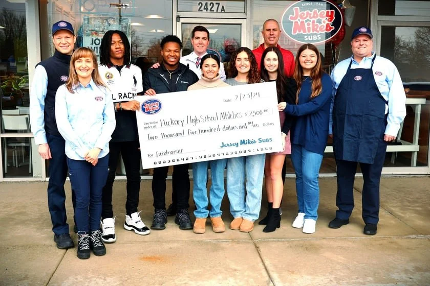 Group photo outside Jersey Mike's Subs featuring people holding a large donation check for Hickory High School Athletics.