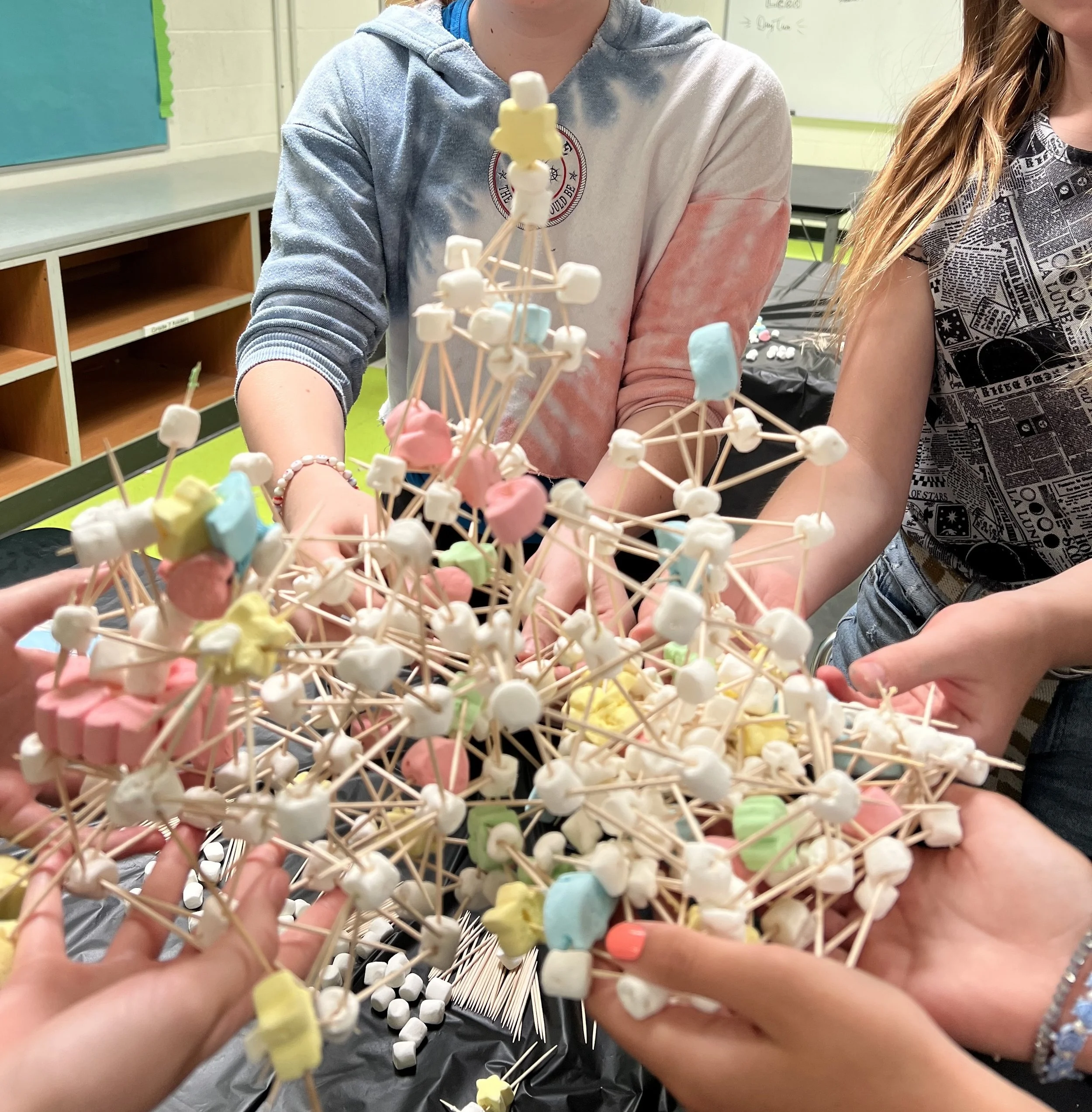 Multiple people assembling a large structure made of marshmallows and toothpicks, with colorful marshmallows in a classroom setting.